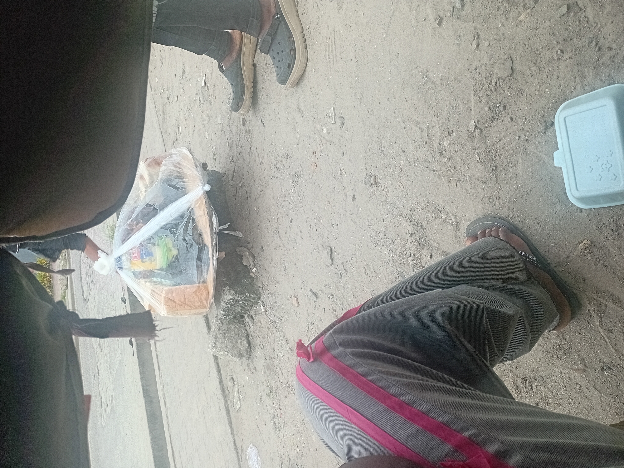 The image depicts an outdoor scene in an urban environment, likely on a street or roadside. In the foreground, a person's legs and feet are visible, wearing grey pants with red stripes and sandals. To the right, another person is seen wearing dark pants and dark clogs. In the middle ground, a plastic bag filled with items, including loaves of bread and a yellow bottle, rests on what appears to be a makeshift platform on the ground. The ground is covered with dirt and small stones. In the background, a paved surface, possibly a road or sidewalk, is visible. The overall scene suggests a street vendor or a roadside gathering. The location is identified as Unknown, Unknown.