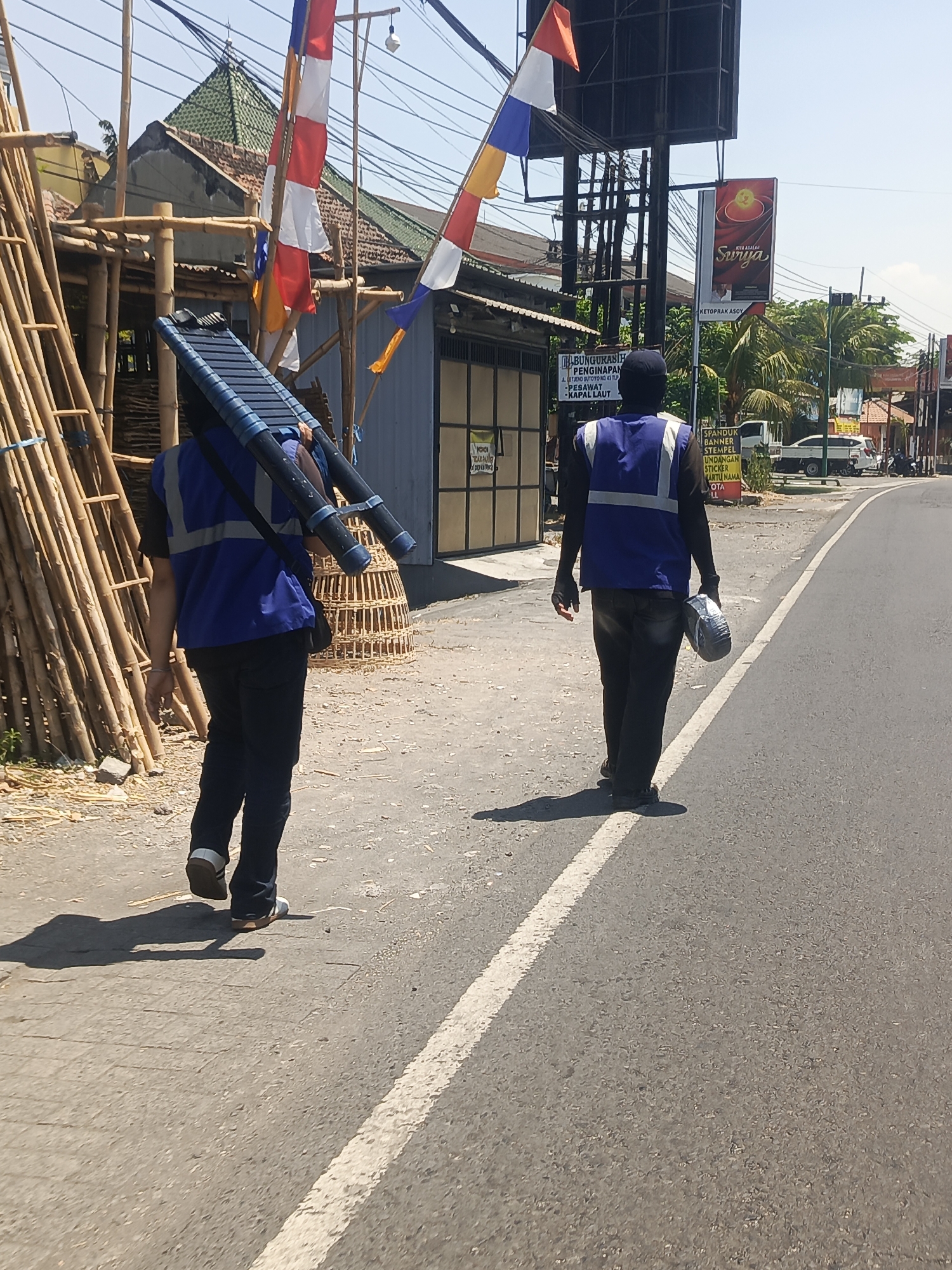 Two individuals are walking away from the camera along a paved road in Kedungrejo, Indonesia, on a bright, sunny day. The individual on the left is carrying a long, dark blue object with handles over their shoulders, possibly a ladder or a tool. They are wearing a blue vest with reflective strips over a dark shirt and dark pants. The individual on the right is carrying a white, cylindrical object in their right hand and is wearing a similar blue vest with reflective strips over a dark long-sleeved shirt and dark pants. In the background, there are buildings, a large billboard advertising "Surga," bamboo scaffolding, and several flags including red and white Indonesian flags. The environment appears to be a semi-urban or rural area with vegetation and trees visible.