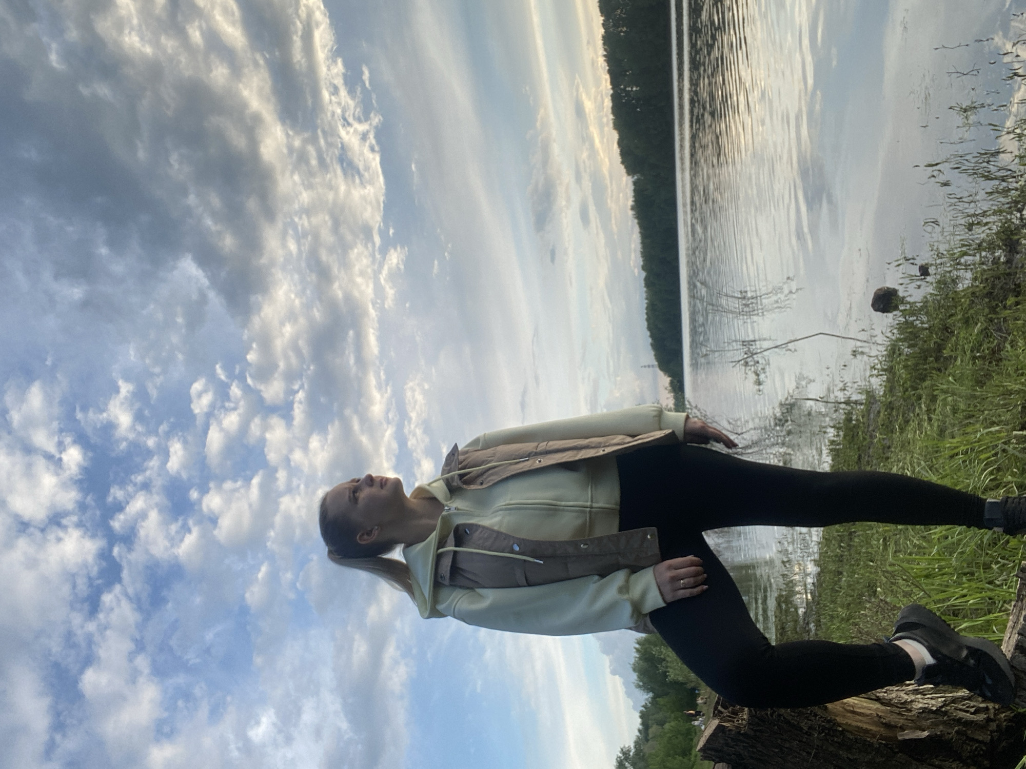 The image depicts a woman posing outdoors. She is wearing black leggings, a light yellow hoodie with a brown vest, and black shoes. She stands on a tree stump with a lake and forest in the background. The sky is filled with clouds. The location is Chaikovsky, Russia.