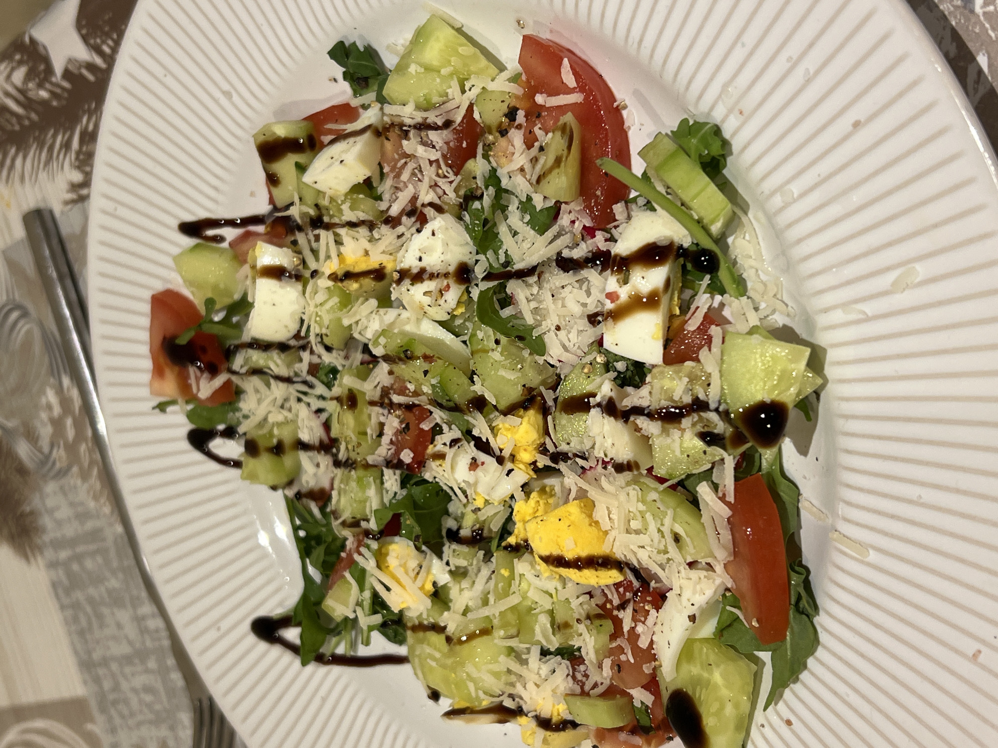 The image shows a close-up, overhead view of a white, ridged plate of salad. The salad consists of arugula, chopped tomatoes, cucumber slices, cubed mozzarella cheese, and hard-boiled egg wedges. It is topped with shredded Parmesan cheese and drizzled with a dark balsamic glaze. The salad is served on a table with a patterned placemat and a silver fork visible in the background. The setting appears to be indoors, possibly a restaurant or dining room, and the lighting suggests it could be evening. There are no people visible in the image. The location is not identifiable from the visual information provided in the image itself; therefore, it is not possible to determine if it is in Balbriggan, Ireland.