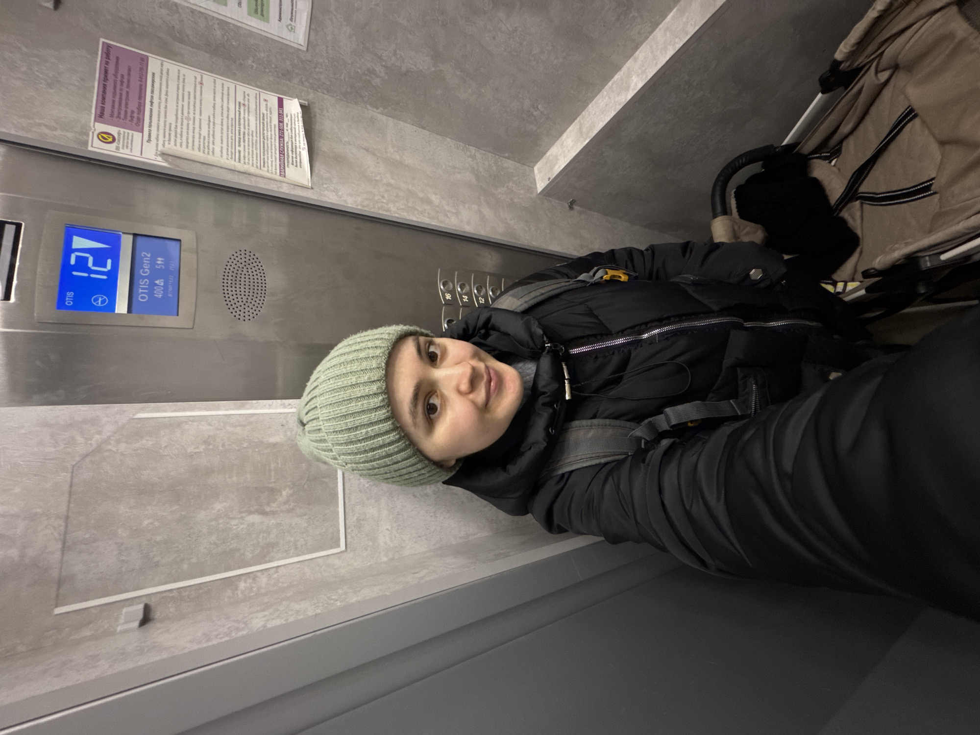 A woman wearing a green knit hat, black jacket, and carrying a backpack is riding in an Otis elevator in Izhevsk, Russia. She is smiling at the camera. The elevator is currently on the 12th floor. A stroller with a beige and black striped lining is also in the elevator. Elevator signs are visible, including passenger instructions, company information, and emergency contact information. The elevator has a gray tiled interior with metal trim.