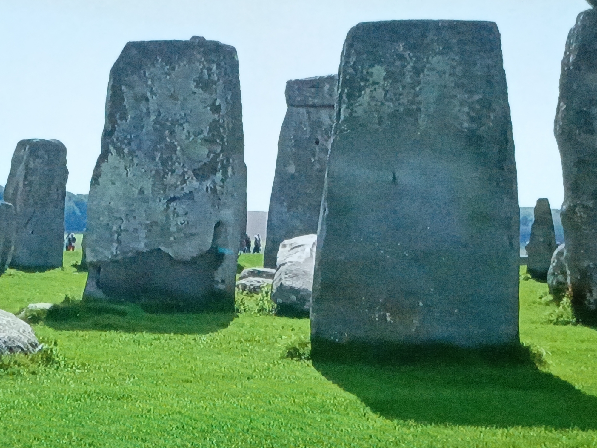 The image shows several large, grey stones standing on a green grassy field under a light blue sky. There are additional stones and figures in the background. This image is not located in Juan Griego, Venezuela. The image depicts Stonehenge.