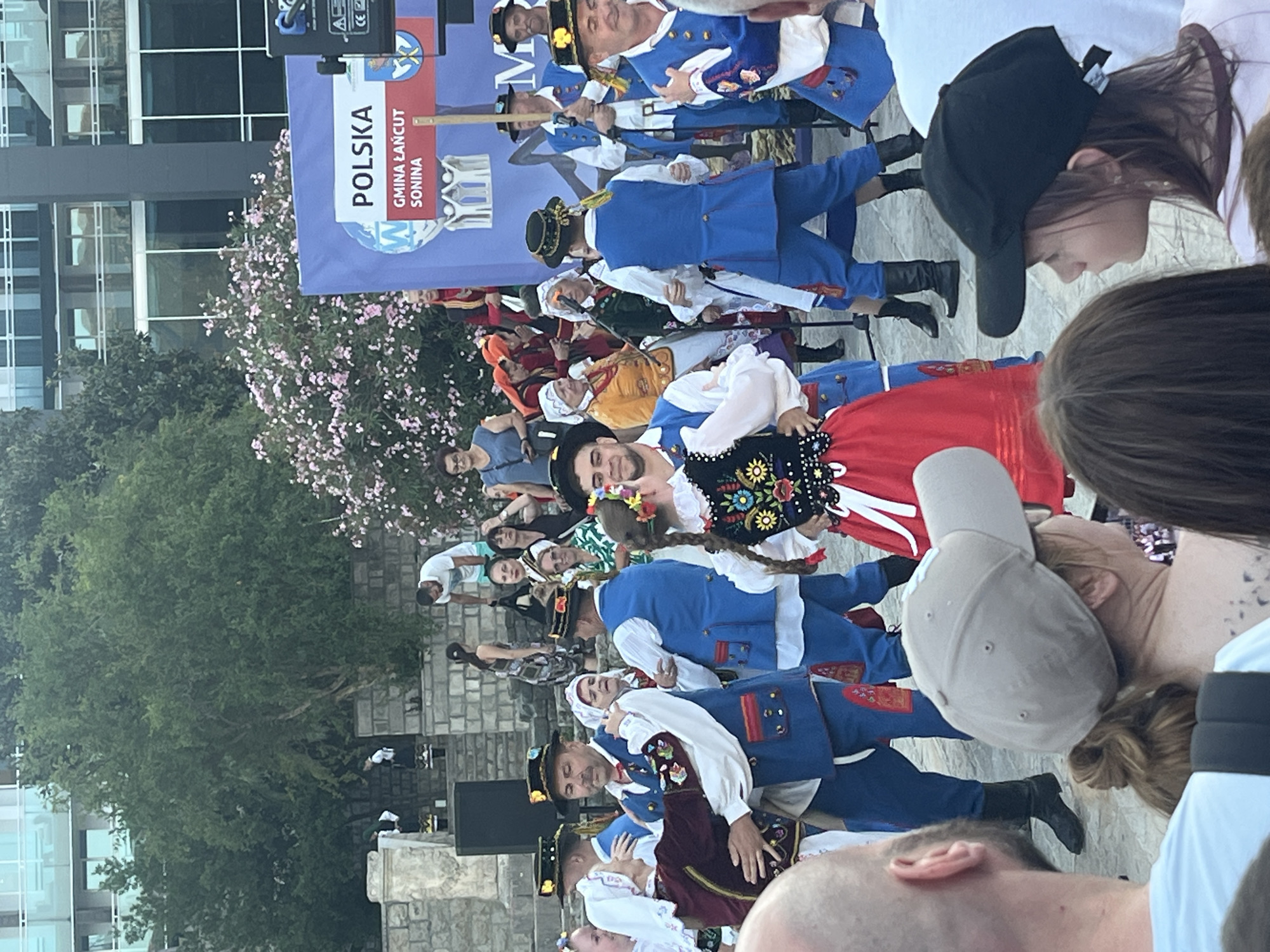 This is a photograph taken outdoors, likely in Budva, Montenegro, given the context. The scene captures a cultural performance, possibly a folk dance, with several individuals dressed in traditional Polish attire. The foreground is partially occupied by the heads of spectators, one wearing a light-colored baseball cap and another a black baseball cap.
In the mid-ground, a group of performers is engaged in dancing. Men are seen in blue vests, white shirts, and trousers, many wearing hats with decorative elements. Some of the men are dancing with women who are wearing colorful skirts and embroidered tops. A woman in a distinctive red skirt with white trim is prominent. Behind the performers, a large blue banner with "POLSKA" and "GMINA ŁAŃCUT SONINA" written on it is visible, indicating a Polish connection. The background features trees with pink blossoms and a modern building with glass windows. The setting appears to be an open area with stone steps and what looks like a stage or performance space. The overall atmosphere suggests a vibrant cultural event.