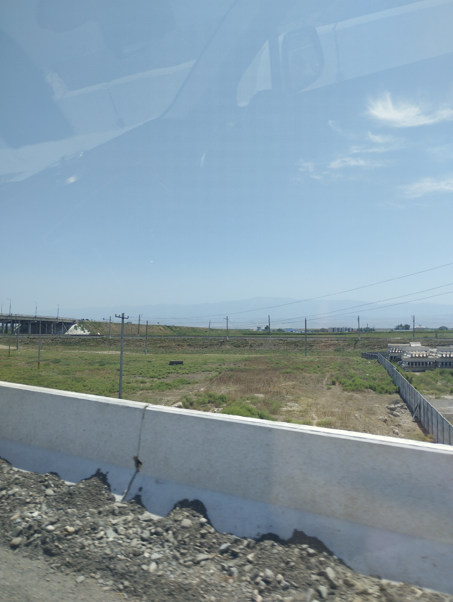 The image shows a view from inside a vehicle, looking out at a landscape under a clear blue sky. In the foreground, a concrete barrier runs horizontally across the bottom of the frame, with a rough pile of gravel and dirt in front of it. Beyond the barrier, a field of dry grass and sparse green vegetation stretches towards a distant landscape. A paved road or railway line is visible in the middle ground, with power lines running parallel to it. Further in the distance, a large bridge or overpass is partially visible on the left. The scene appears to be an undeveloped or rural area, possibly on the outskirts of a city. The environment suggests a warm, dry climate. There are no people or animals visible in the image. The overall impression is one of open space and infrastructure development in a semi-arid region.