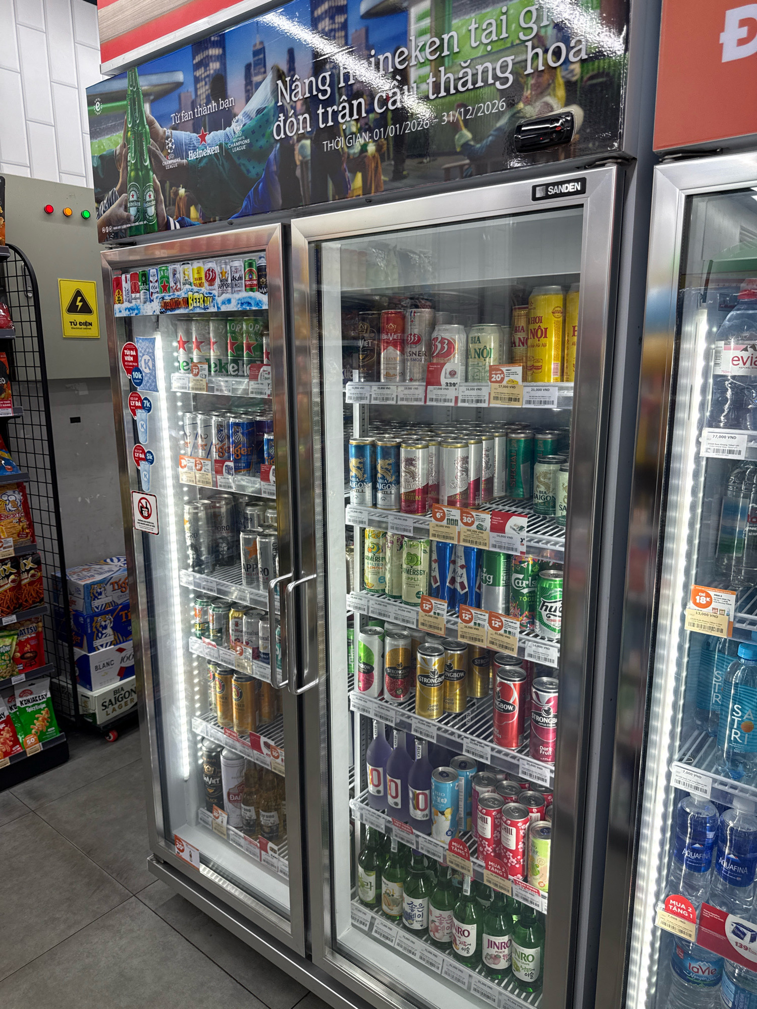 This is a medium-shot, eye-level, exterior photograph of a commercial refrigerator stocked with beverages, likely taken inside a convenience store or supermarket. The refrigerator has two glass doors, one of which is slightly ajar, revealing rows of canned and bottled drinks. The top of the refrigerator is adorned with a large promotional banner featuring a Heineken beer advertisement in Vietnamese.
The promotional banner displays text such as "Nâng ly thành bạn," "đón trận cầu thăng hoa," "THỜI GIAN: 01/01/2026 - 31/12/2026." The banner also includes images of Heineken beer bottles and a soccer stadium. To the left of the refrigerator, a yellow warning sign with a lightning bolt symbol and Vietnamese text "TỦ ĐIỆN" (electrical cabinet) is visible, along with three colored indicator lights (red, orange, green). Several racks of snacks and other products are also visible to the left and on top of the refrigerator.
The drinks inside the refrigerator include various brands of beer and other beverages in cans and bottles. Some visible labels include "Heineken," "Bia Saigon," "Tiger," "Sapporo," "Strongbow," and "Jinro." The refrigerator is illuminated by internal LED lighting.
The floor is tiled with large, light-colored square tiles. The overall environment suggests a retail setting. The presence of Vietnamese text on the signage and product labels indicates the location is in Vietnam. Given the context of a convenience store chain like Circle K or similar, and the style of advertisements, it is highly probable this image was taken in Nha Trang, Vietnam, a coastal city known for its tourist attractions and numerous convenience stores catering to both locals and tourists.
