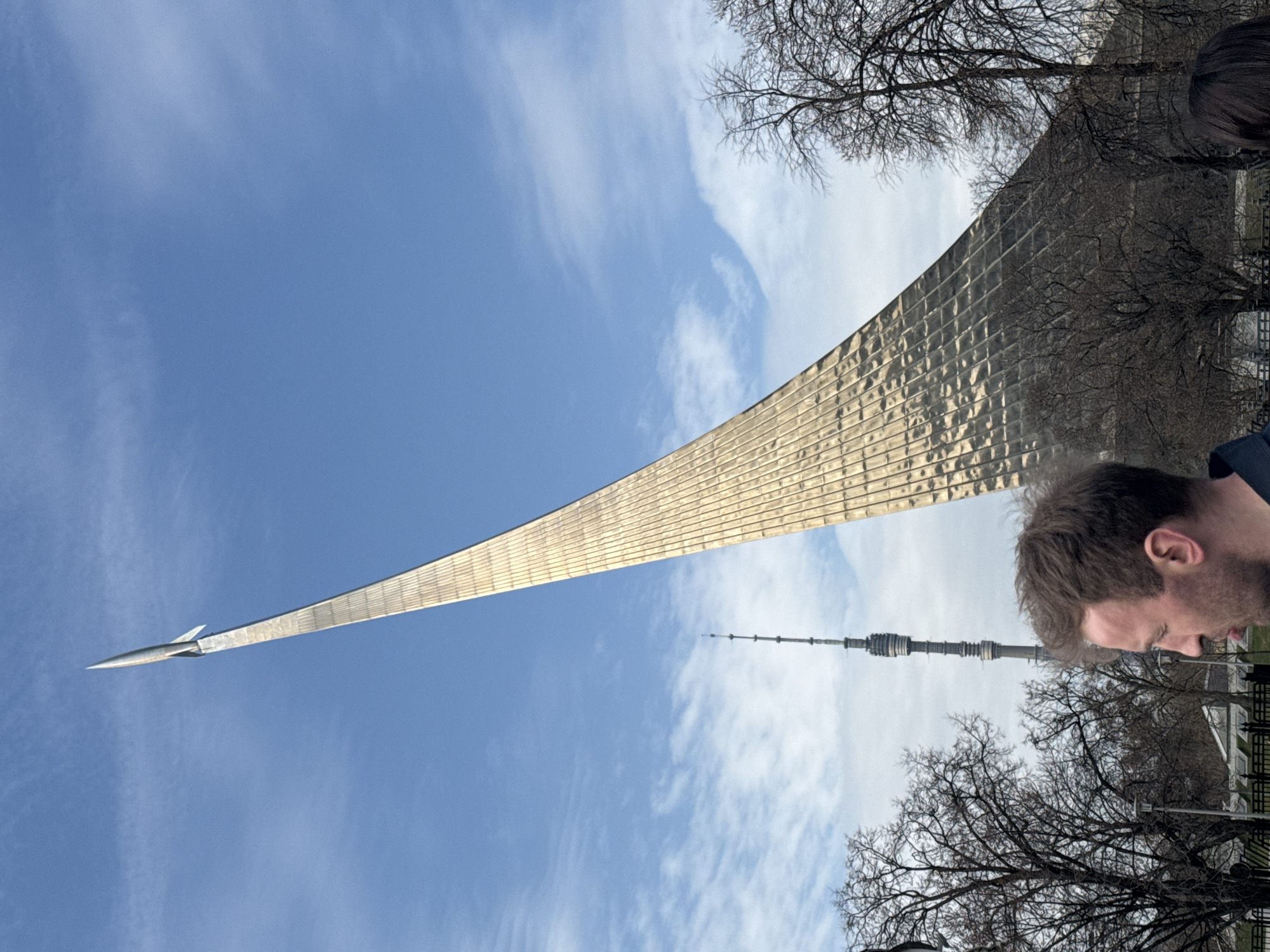 The image captures the Monument to the Conquerors of Space in Moscow, Russia, set against a partly cloudy sky. The monument is a tall, curved structure covered in metallic tiles, topped with a model rocket. Adjacent to it, and partially obscured by bare trees, is the Ostankino Tower. A man with short brown hair is visible in the foreground, seemingly observing the scene. The environment appears to be a park or similar public space, evidenced by a railing.
