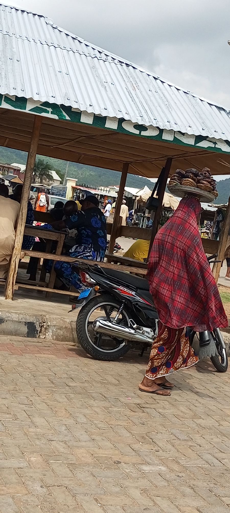 A woman in Lokoja, Nigeria, wearing a red plaid shawl and patterned skirt, is walking on a brick-paved area with a flat round tray of food items balanced on her head. The woman is wearing brown sandals. In the background is an open-air structure with a white corrugated metal roof, supported by wooden beams. There are people sitting and standing inside the structure. A black motorcycle is parked next to the structure. Hills are visible in the distance.