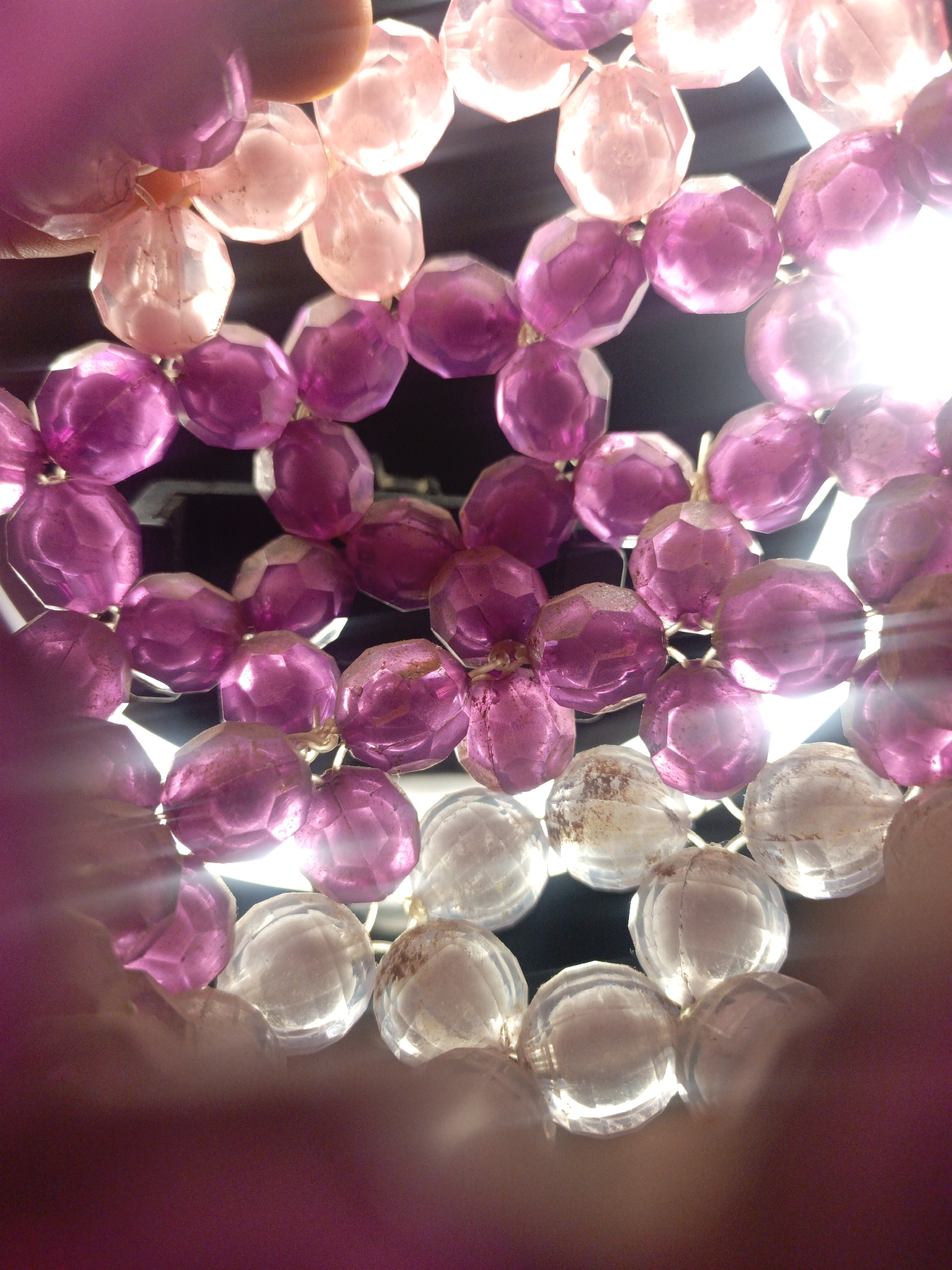 A low-angle, close-up shot captures a strand of faceted beads, some translucent pink and some a deep amethyst color, against a dark background. The beads are strung together and appear to be part of a larger decorative item, possibly a garland or a section of jewelry. The lighting is bright and appears to be coming from behind the beads, creating highlights and a lens flare effect that obscures some of the details. A person's hand, also a soft pinkish hue, is visible in the upper left, gently holding the beads. The overall impression is one of delicate craftsmanship and vibrant color. The location is unknown.