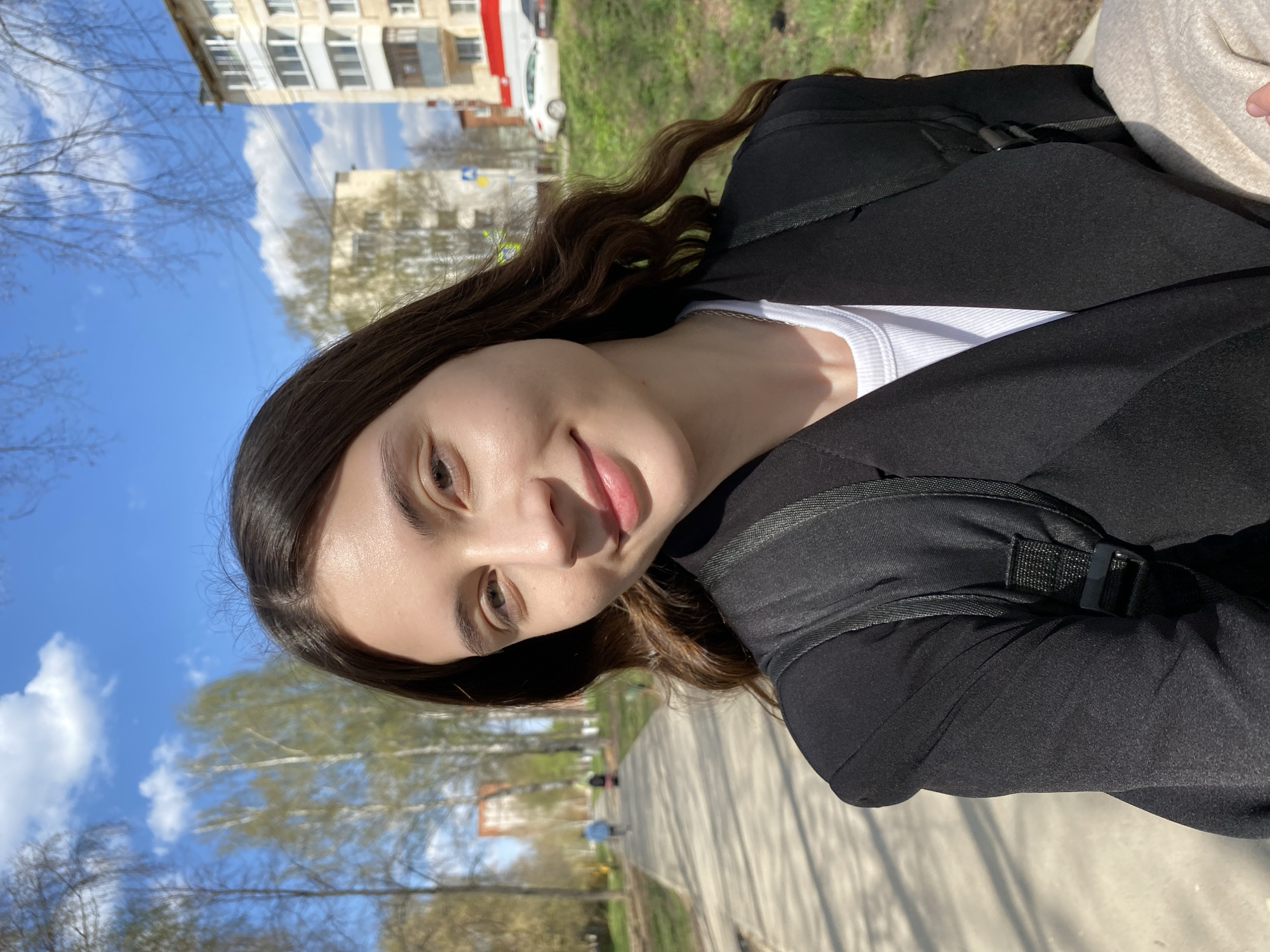 A young woman is outdoors in Votkinsk, Russia. She wears a black jacket over a white top, and a backpack. Her long brown hair is wavy. She's on a paved path, with trees and grass visible on the side. Buildings are in the background, one appears to be residential. The sky is blue with clouds. The lighting suggests it is daytime.