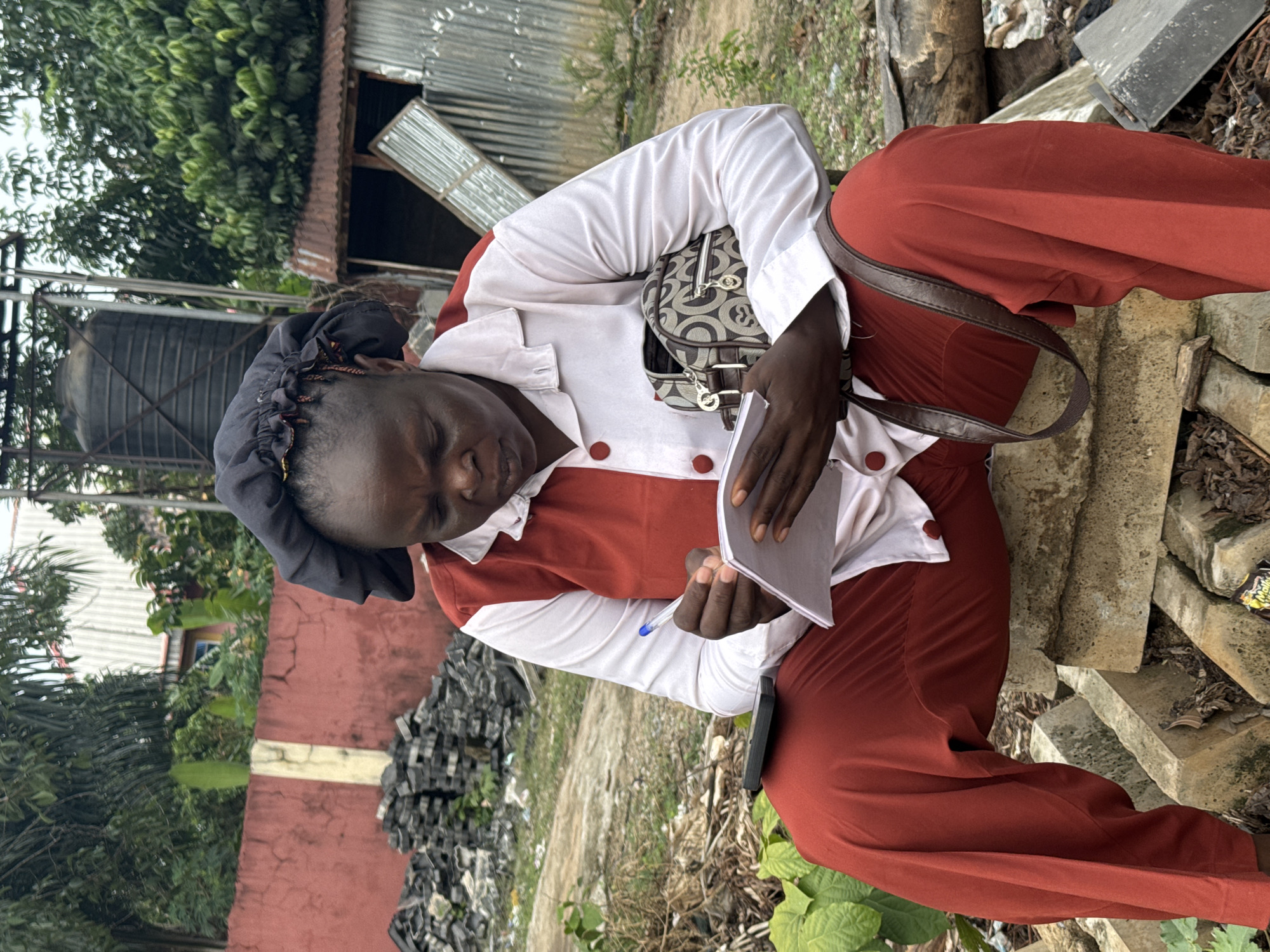 In Orifite, Nigeria, a woman wearing red pants, a white shirt with red trim, and a black head covering sits outdoors, hunched over a notebook and pen. She has a patterned handbag slung over her shoulder. She is positioned on what appears to be concrete blocks or debris. Behind her are scraps of metal and other debris. A water tank and buildings are visible in the background. There are also trees and plants.