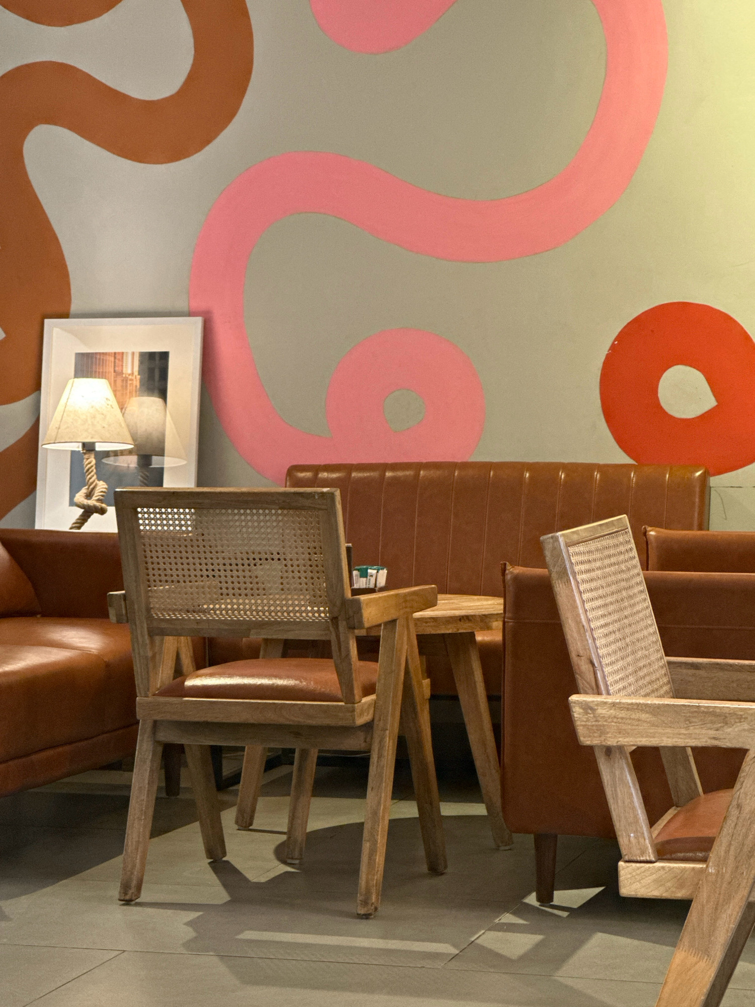 The image captures a seating area within a cafe in Lalitpur, Nepal. In the foreground, two wooden chairs with cane-woven backs and leather seats are positioned near a small wooden table. A brown leather sofa is visible to the left and right, and a longer brown leather banquette seating extends across the back of the scene. A framed picture, depicting a cityscape with a lamp, hangs on the wall behind the sofa on the left. The wall itself is a muted gray, adorned with large, abstract swirls of pink and orange paint. The floor appears to be tiled in a neutral tone. There are no people present in the image.