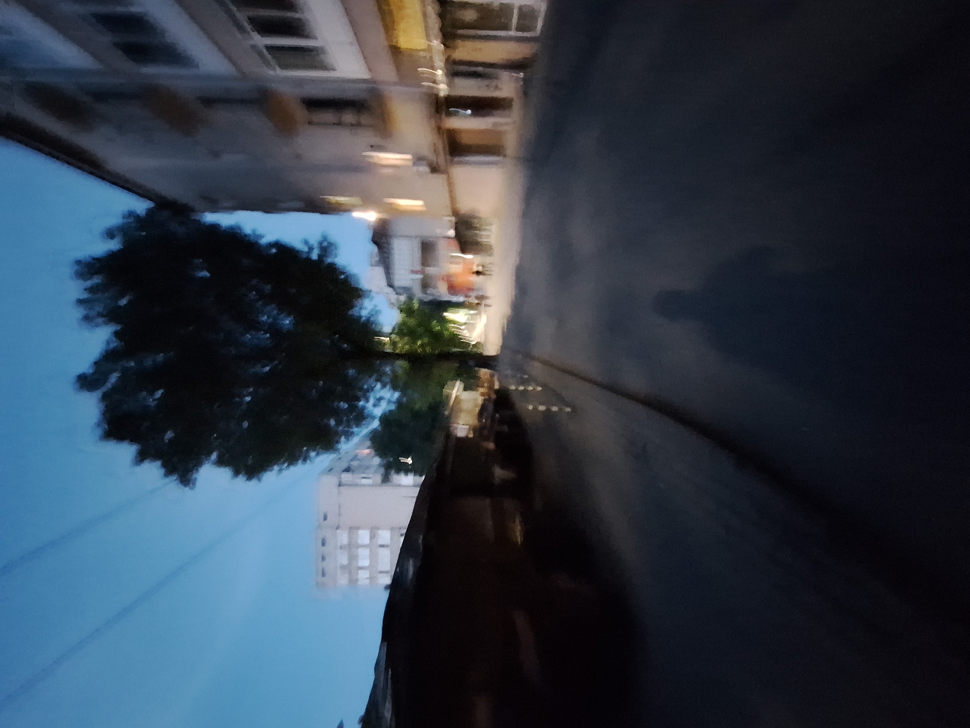 The image depicts a street scene in Varna, Bulgaria, likely in the evening or early morning given the low light conditions and the blue hue of the sky. In the foreground, the asphalt road is visible, with a shadow of a person, possibly the photographer, cast upon it. To the left, a dark, low-lying structure with a sloped roof occupies a significant portion of the frame. Further back, a large tree with dark foliage stands prominently. On the right side of the street, buildings line the sidewalk. The building closest to the foreground has multiple windows and some illuminated interior lights. Beyond this, other buildings with varying architectural styles are visible, some appearing taller and more modern. A few indistinct figures can be seen walking or standing on the sidewalk further down the street, suggesting pedestrian activity. Power lines are faintly visible against the sky. The overall impression is of a quiet urban environment at dusk.