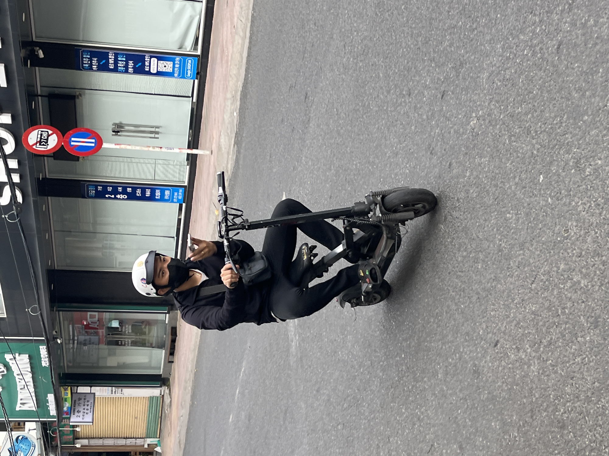A person wearing a white helmet and a black face mask is riding a black electric scooter on a paved road in Nha Trang, Vietnam. The individual is dressed in black clothing, including a hooded jacket and pants, and has a small black bag slung across their chest. They are holding a mobile phone to their ear with their right hand while steering the scooter with their left hand. The background shows the exterior of a commercial building with glass windows and signage. A street sign with red and white circular symbols is visible on the right side of the image. The road surface is asphalt with visible cracks and debris. The overall scene depicts an everyday urban streetscape.