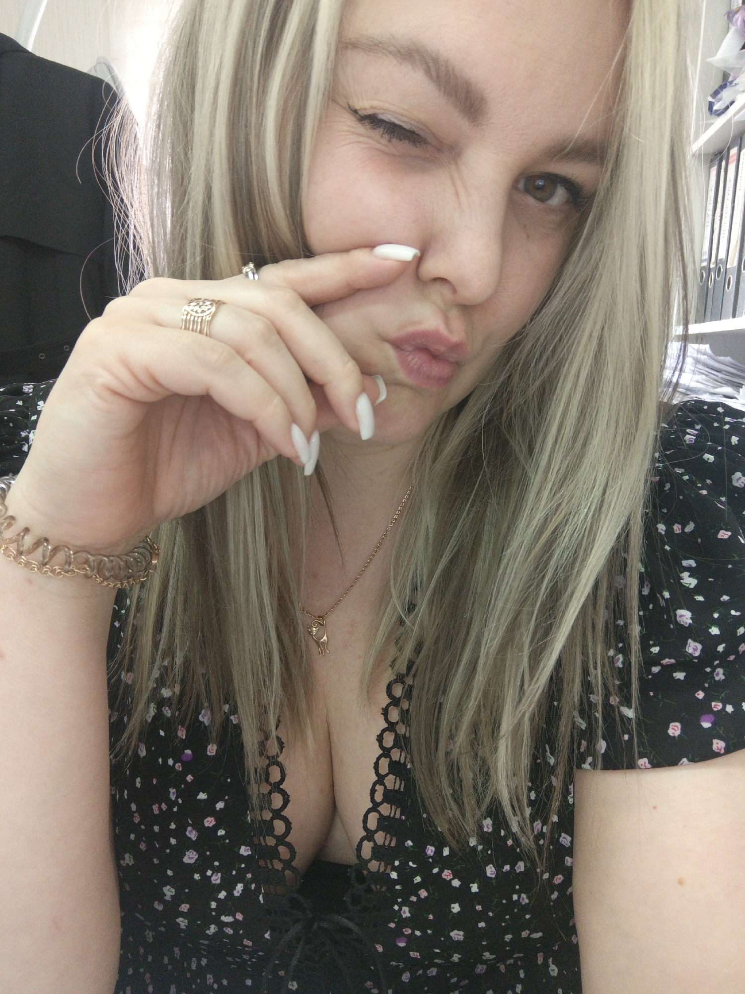 A woman with blonde hair with streaks of lighter blonde is winking and holding her nose with her fingers while pouting her lips. She is wearing a black floral dress with a low neckline, a gold bracelet, a gold ring, and a gold necklace with a cat pendant. Her nails are long and white. In the background, there is a black office chair and a shelf with binders. The location cannot be determined from the given image.