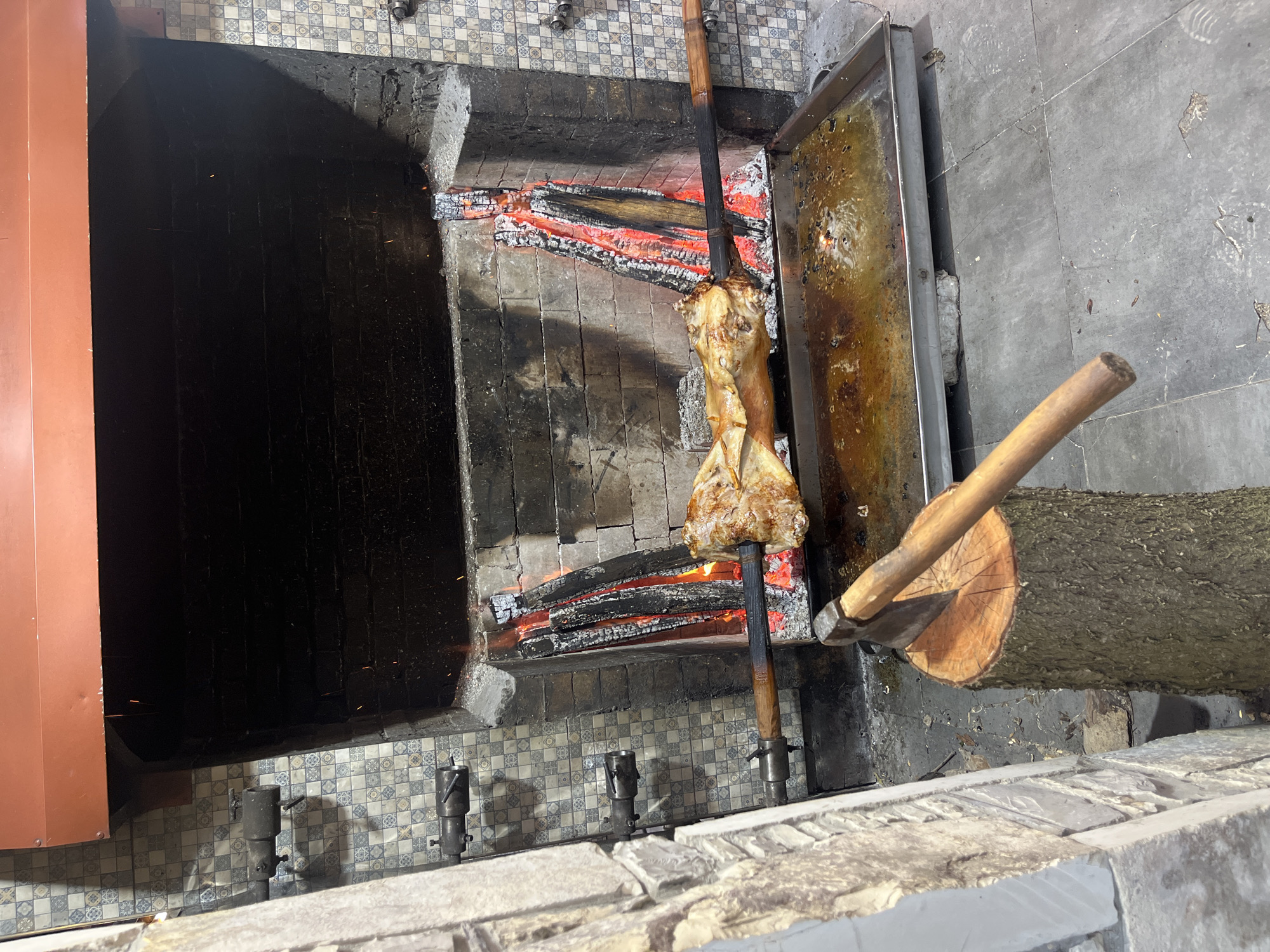 A piece of meat is being cooked over a fire in a large indoor grill. The meat, on a vertical wooden skewer, is positioned over hot coals within a brick and tile-lined structure. To the right of the grill, a metal tray is positioned to catch drippings, next to a large log with a partially embedded axe. The setting appears to be a kitchen or restaurant. The location in Miatli, Russia is provided as external information.
