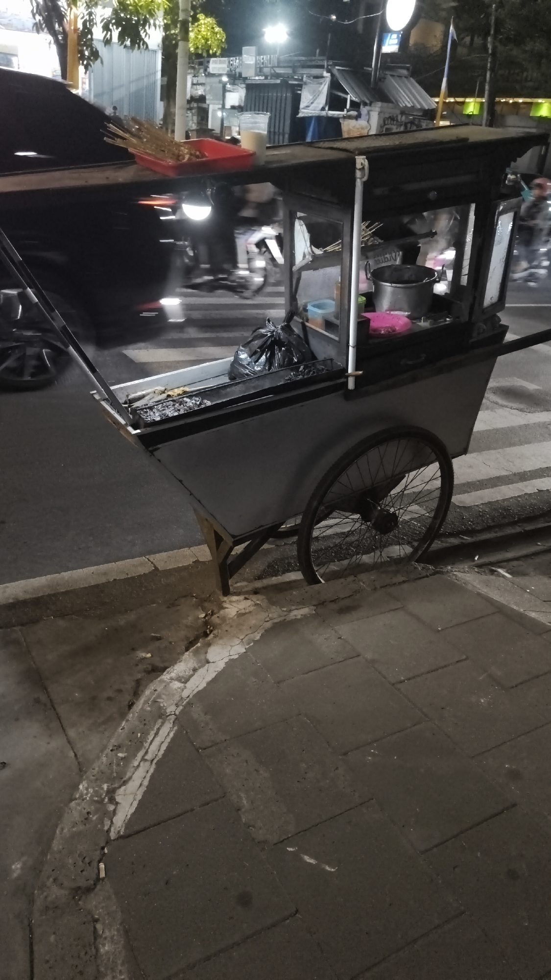 The image depicts a food cart at street level, likely in South Jakarta, Indonesia, at night. The cart, made of dark metal and wood, is positioned on a sidewalk adjacent to a street. A large, black trash bag is visible inside the cart, and a red plastic container holding skewers of food sits on the cart's upper surface. A metal grill is also present within the cart. A spoked wheel is attached to the cart. In the background, other food stalls or businesses are dimly illuminated, and motorcycles are passing on the street, their headlights creating streaks of light. The sidewalk is paved with square tiles and shows signs of wear and cracking. A black car is parked or driving by on the left side of the frame.