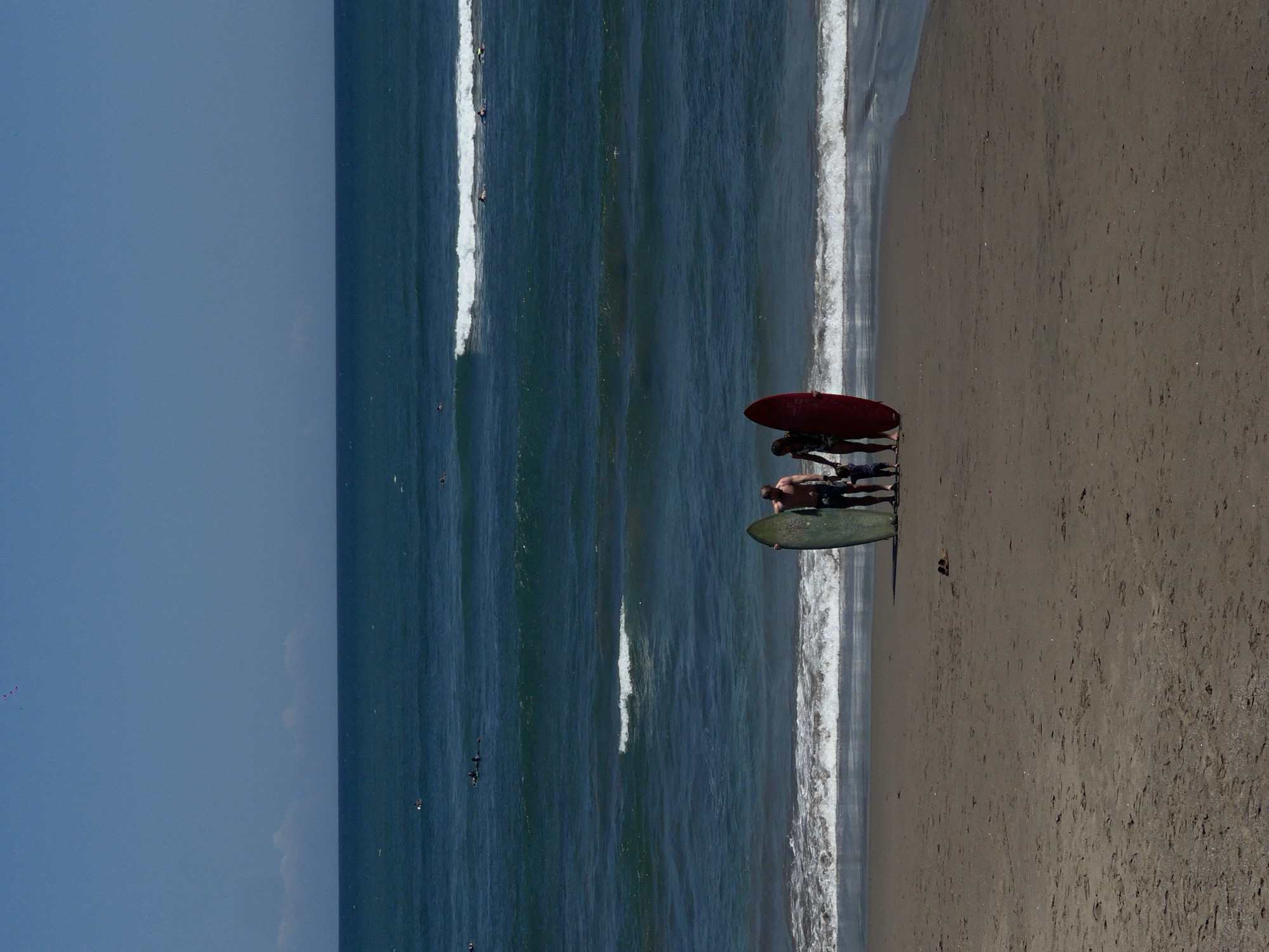 The image shows a group of people with surfboards on the beach at North Kuta, Indonesia. There are two surfboards visible, one red and one green. There are multiple people visible. The ocean is present with waves breaking on the shoreline. The sky is clear.