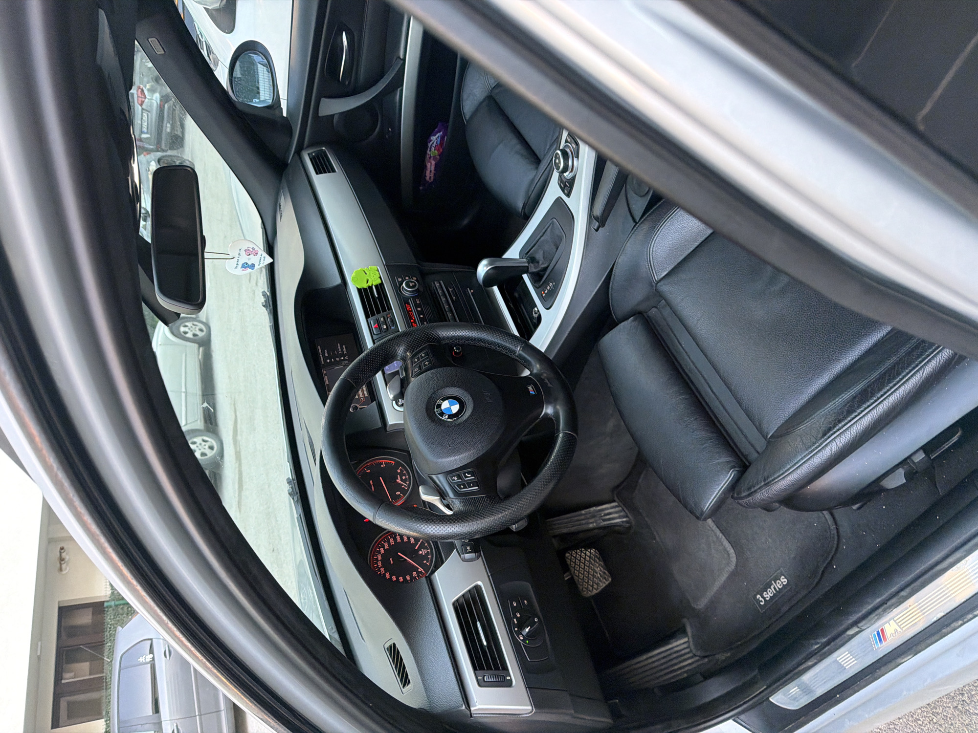 The image shows the interior of a BMW 3 Series, taken from the passenger side. The steering wheel, dashboard, and front seats are visible. A rearview mirror is present, and objects can be seen through the car's windows. The car is likely in Sofia, Bulgaria, based on the presence of Bulgarian language elements in the image (e.g. a heart-shaped object hanging from the rearview mirror).