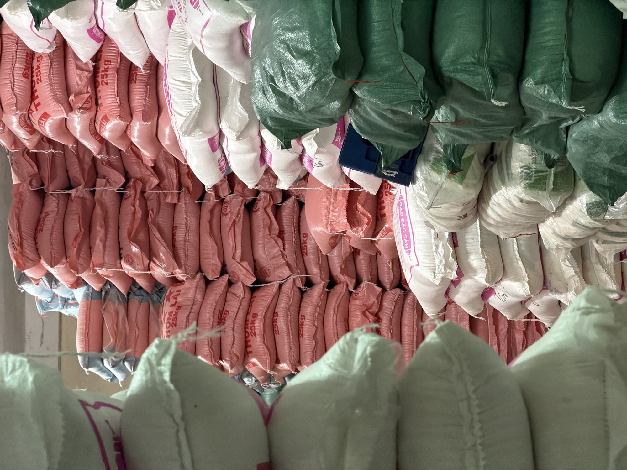 The image shows rows of stacked sacks, likely filled with rice, hanging and resting on the floor, suggesting a warehouse or storage facility. The sacks are predominantly pink and white, with a smaller section of green sacks. Some sacks are labeled with "25kg" and "HOM LAI" suggesting their weight and possibly the type of rice. The background is blurred, obscuring details of the location. Đà Nẵng, Vietnam is only relevant to the OCR text of a product shown.