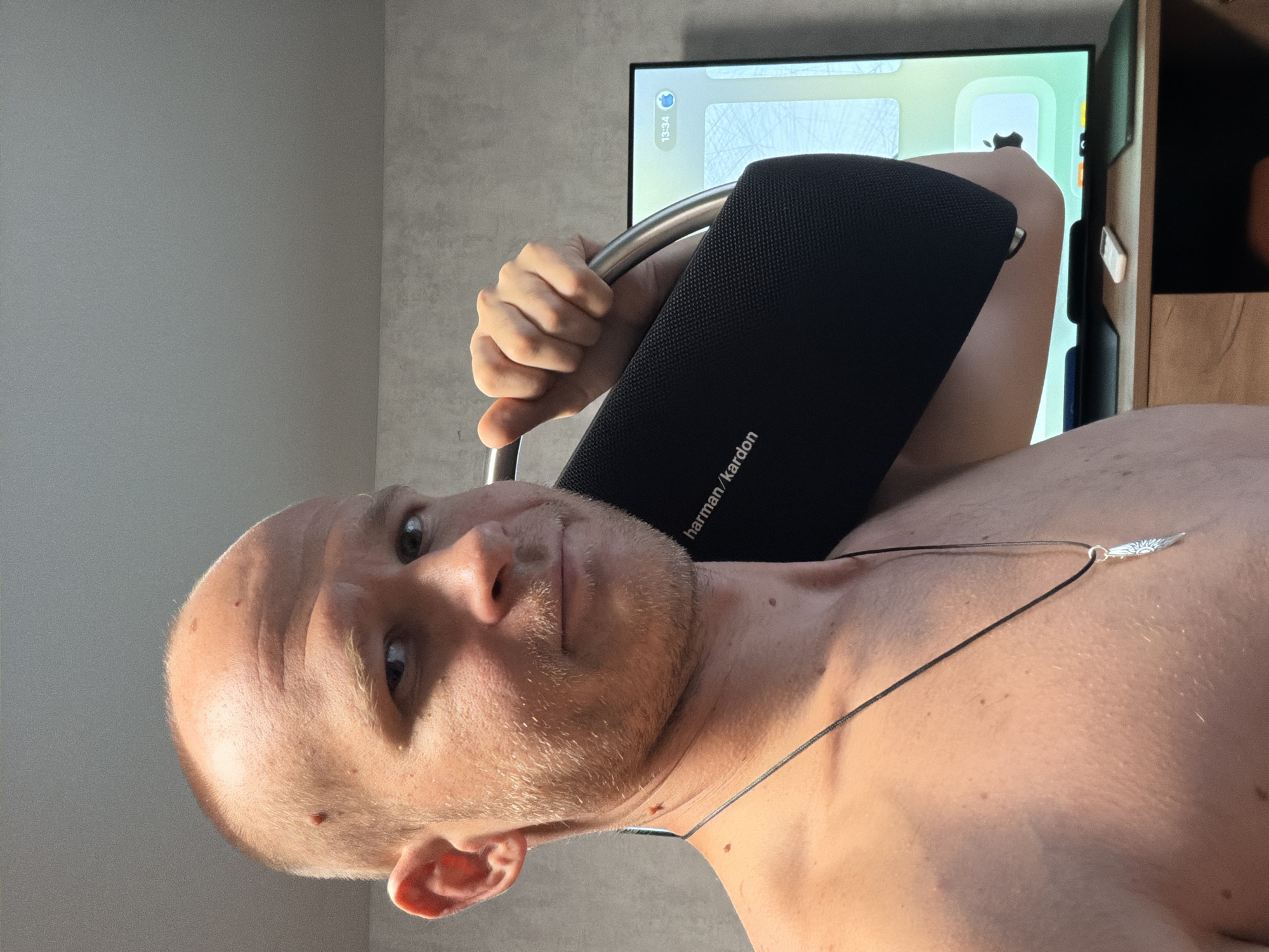 A man with a shaved head and stubble is holding a black Harman Kardon speaker over his shoulder. He is shirtless and wearing a necklace with a silver pendant. A television screen is visible in the background, displaying a time of 13:34 and an Apple logo. The scene appears to be indoors, possibly in an apartment in Yekaterinburg, Russia. The walls are a neutral gray color, and there is a wooden cabinet visible beneath the television. The lighting suggests it is daytime.