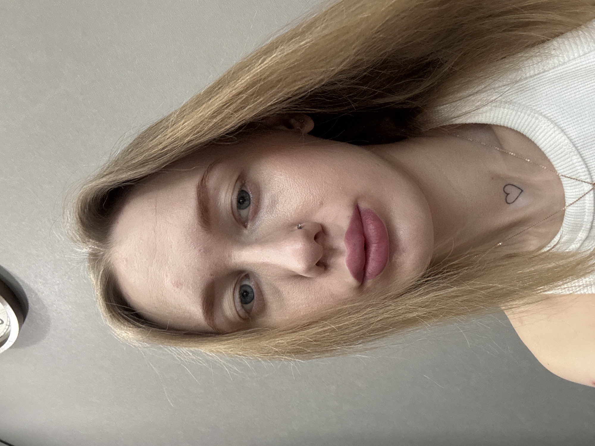A young woman with blonde hair and fair skin is shown in a close-up, indoor photograph. She is facing the camera at an angle. She has light blue eyes, a pierced nose, and full, pink lips. A small heart tattoo is visible on her upper chest. She is wearing a white shirt. The background is a plain, light gray wall. There is no information to conclude the city is in Ovoshchnoy, Russia.