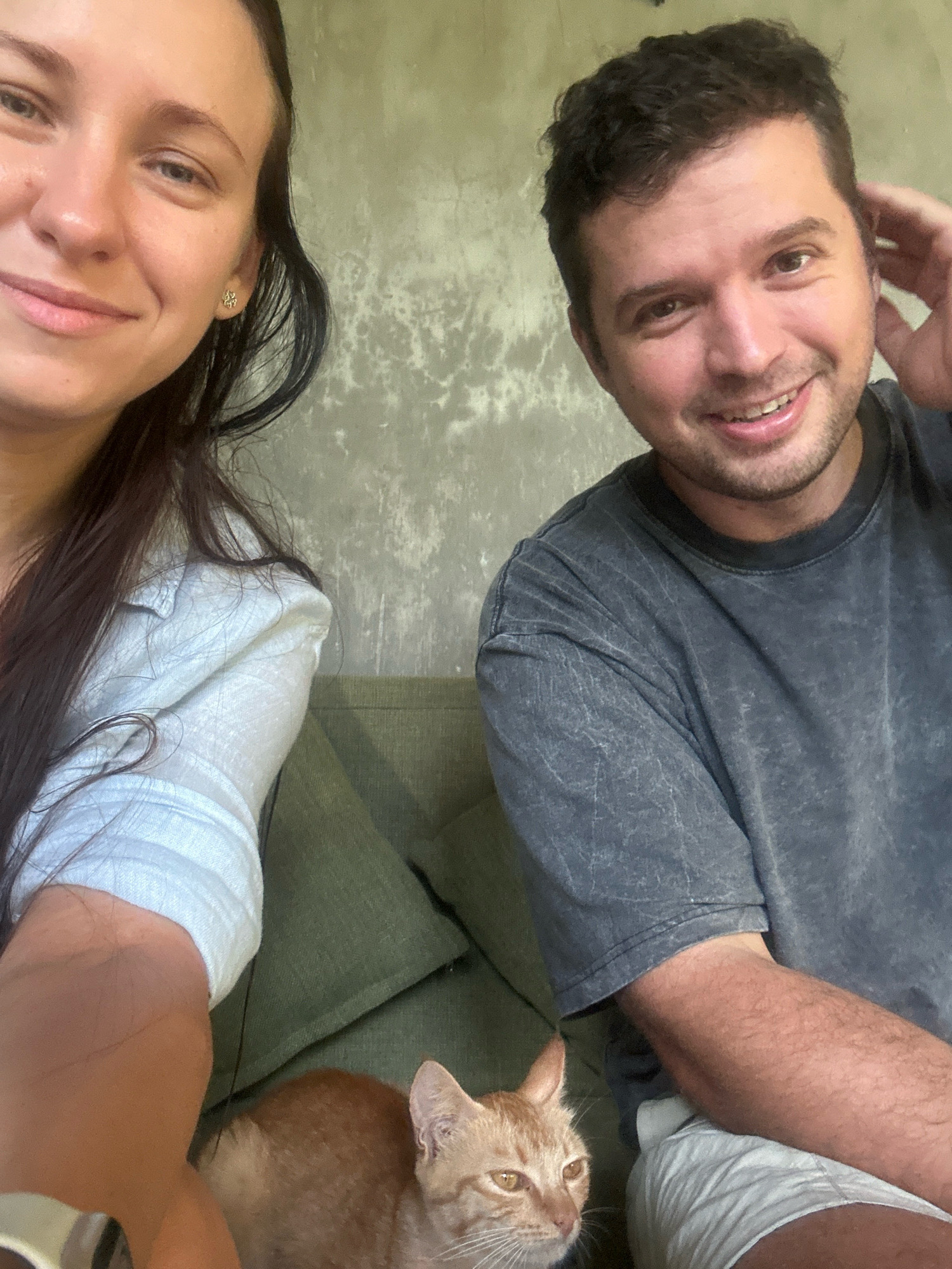 A selfie is taken indoors, likely in a living room, with a man and a woman smiling at the camera. The woman is on the left, wearing a white button-down shirt and a clover-shaped earring. The man is on the right, wearing a dark, distressed t-shirt. Between them, a ginger cat is nestled on a green sofa. The background features a textured gray wall.