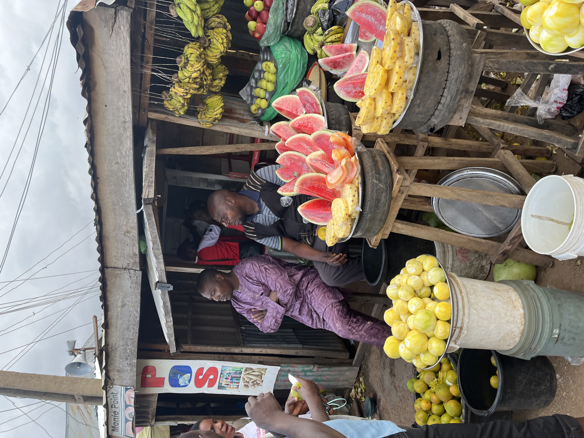 The image shows an outdoor fruit stand in Gura Top, Nigeria. Several men are present, with two seated and one standing, appearing to be engaged in a transaction. The stand displays a variety of fruits, including watermelons, pineapples, and bananas, arranged on trays and in buckets. Overhead, a corrugated metal roof covers the stand, and electrical wires are visible. The surrounding environment suggests a busy marketplace.
