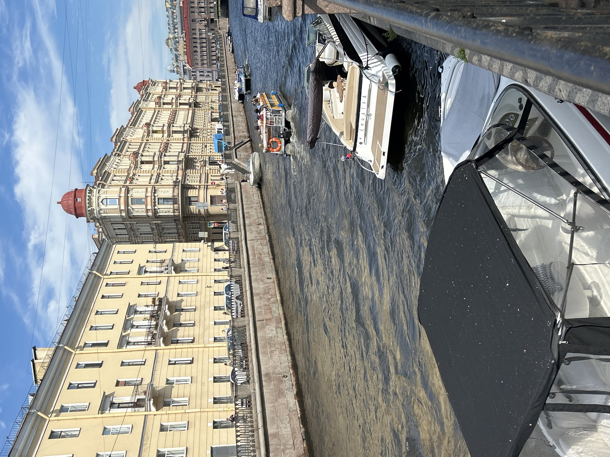 This media file depicts a canal scene in a city, likely during daytime given the bright sunlight casting shadows. The sky is blue with scattered white clouds.
The foreground is dominated by the bow of a white speedboat, partially covered by a black tarp. Reflections of sunlight are visible on the boat's windshield. Further into the canal, another white boat with a beige interior is visible, also appearing to be a speedboat. It has the word "СИНАТРА" (SINATRA in Cyrillic) written on its side in a darker color. A group of people are seated on this boat, suggesting a sightseeing tour or leisure activity. Behind it, a larger yellow tour boat with an orange life preserver is visible, also carrying passengers.
Along the canal's edge, a stone embankment lined with a metal railing separates the water from a building complex. The buildings are a mix of architectural styles. On the left, a tall, pale yellow building with numerous windows and balconies is prominent. To its right and further down, a more ornate building with a red dome and intricate facade details stands out. The buildings suggest an established urban environment, possibly with historical significance.
The water in the canal is dark and appears to be flowing, with ripples and reflections on its surface. The overall impression is a bustling urban waterway on a clear, sunny day, with boats transporting people and iconic architecture forming the backdrop.