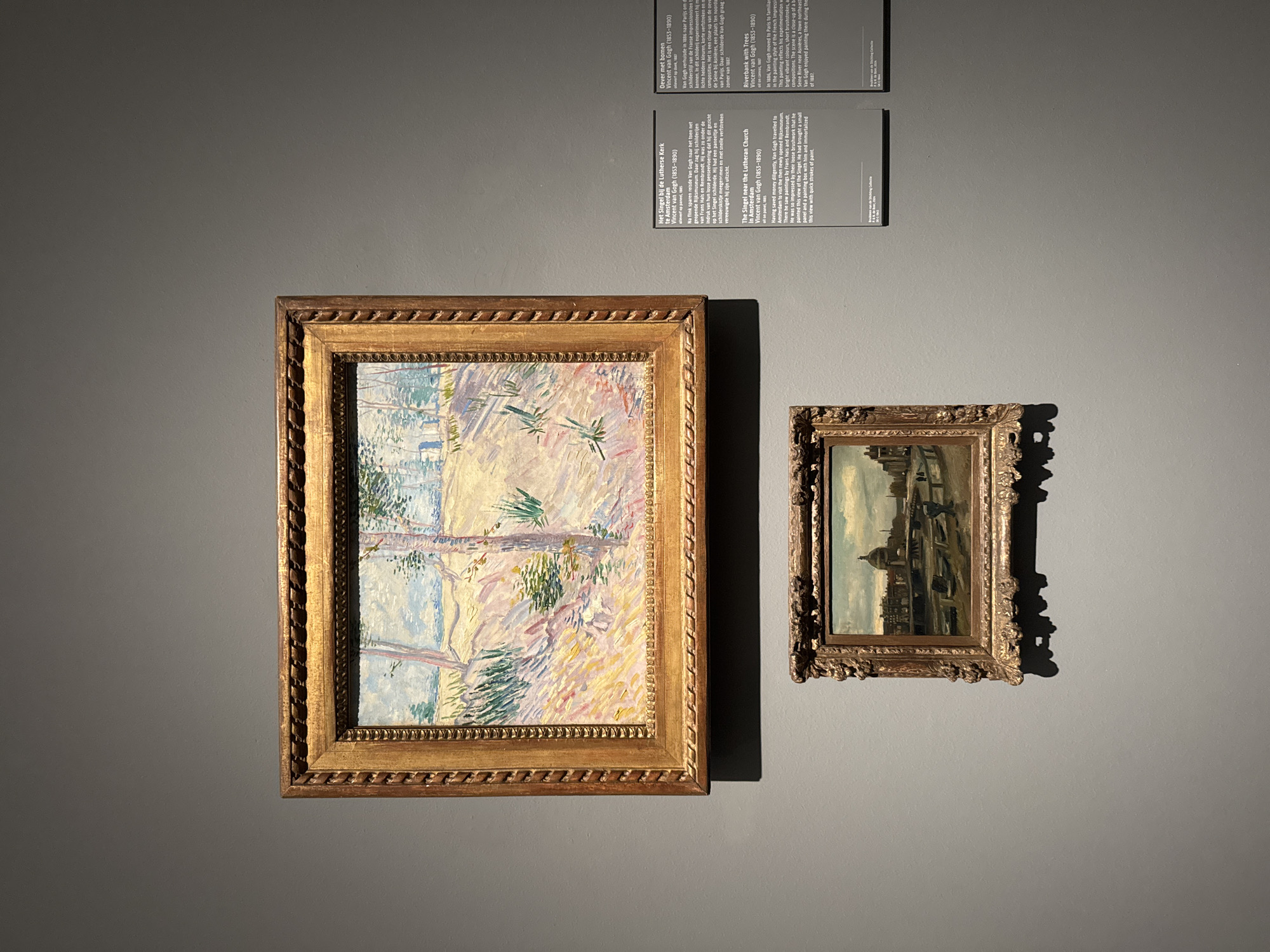 Two framed paintings hang on a gray wall, accompanied by three informational panels. The larger painting, on the left, features a landscape with trees in impressionistic style, with light, bright colors. The painting is in an ornate, gilded frame. The smaller painting, to the right, is also in a gilded frame, and depicts a cityscape, also in an impressionistic style. The informational panels above the paintings contain text in Dutch and English, describing the works and their creator, Vincent van Gogh. The text mentions locations in Amsterdam, including the Singel near the Lutheran Church and the Seine River near Asnières.