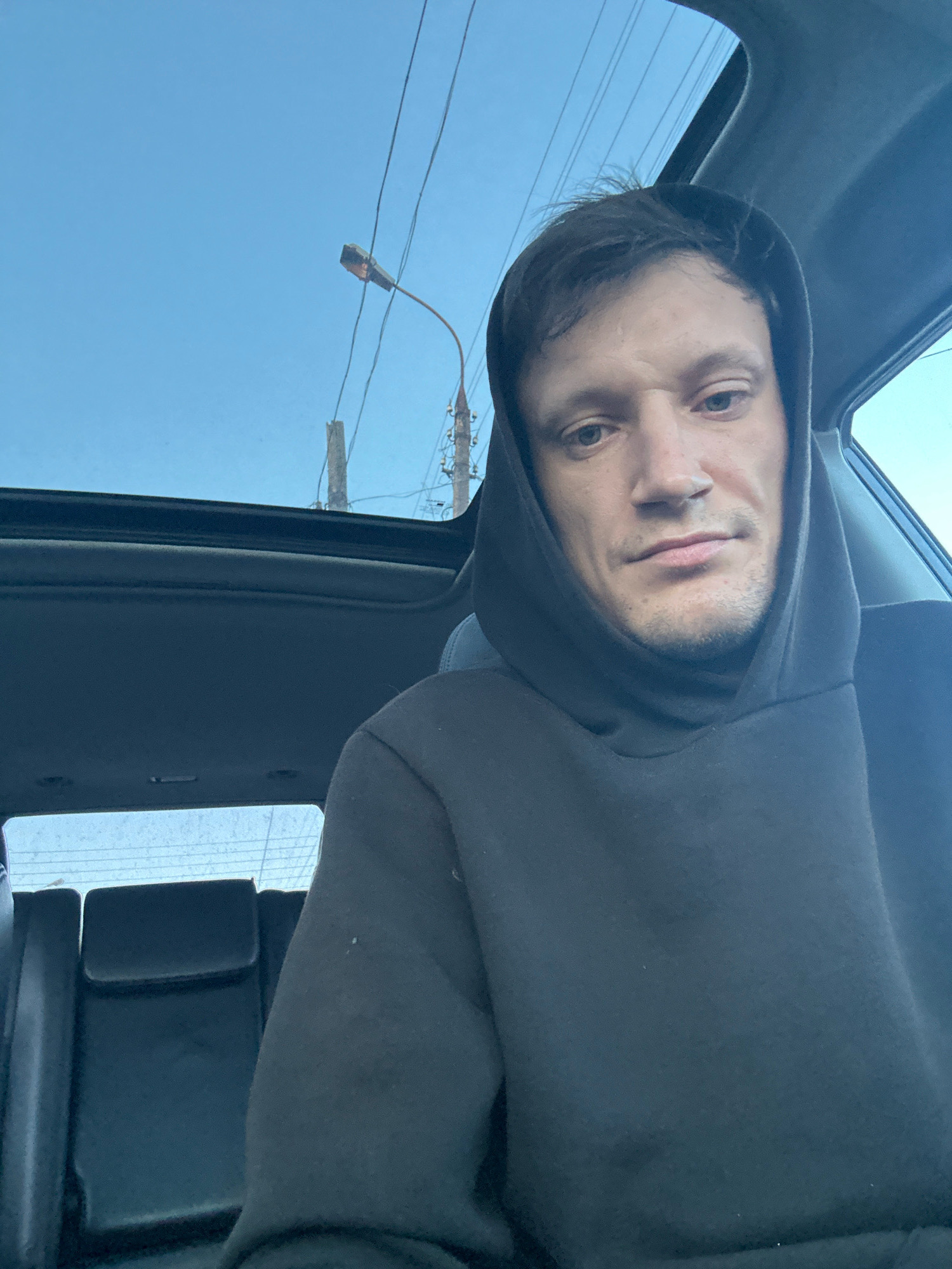 A man in a black hoodie is sitting in the passenger seat of a car, looking at the camera. He has short, dark hair and light-colored eyes. The car has a sunroof, and through it, a bright blue sky with some power lines and a streetlamp are visible. The interior of the car is dark, with black seats in the background. The scene appears to be in Makiv, Ukraine, based on the request.
