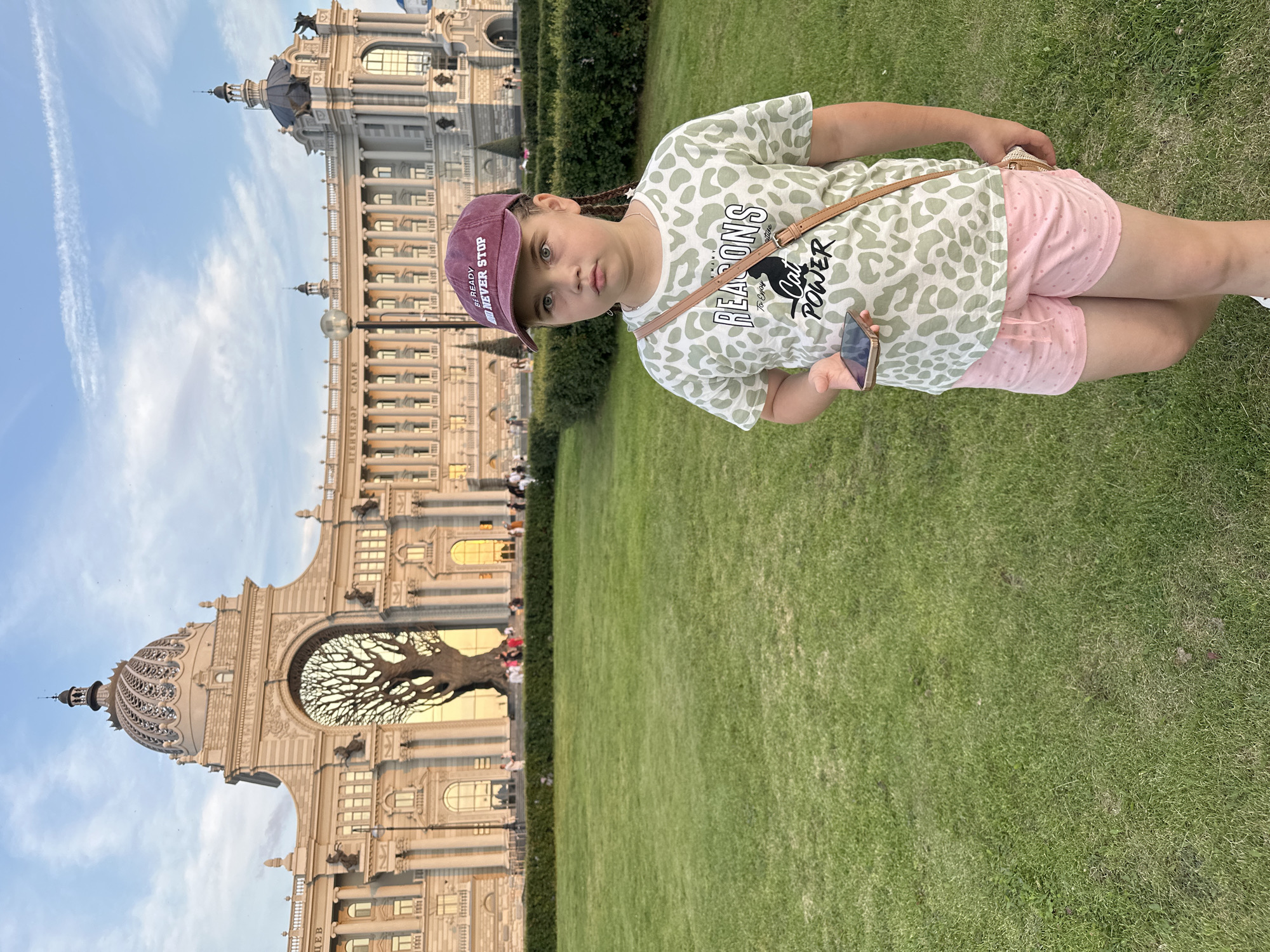 A young girl with braided hair, wearing a maroon baseball cap, a light-colored shirt with a leopard-print and text that says "REASONS To ENJOY Cat POWER", and pink polka-dot shorts, stands in a grassy field in front of a large, ornate building. She is holding a smartphone in her right hand. The building, identified as the Palace of Agriculture in Kazan, Russia, features a grand arched entrance framing a tree. The sky is cloudy and appears to be late afternoon or early evening. Several people are visible in the background near the building.