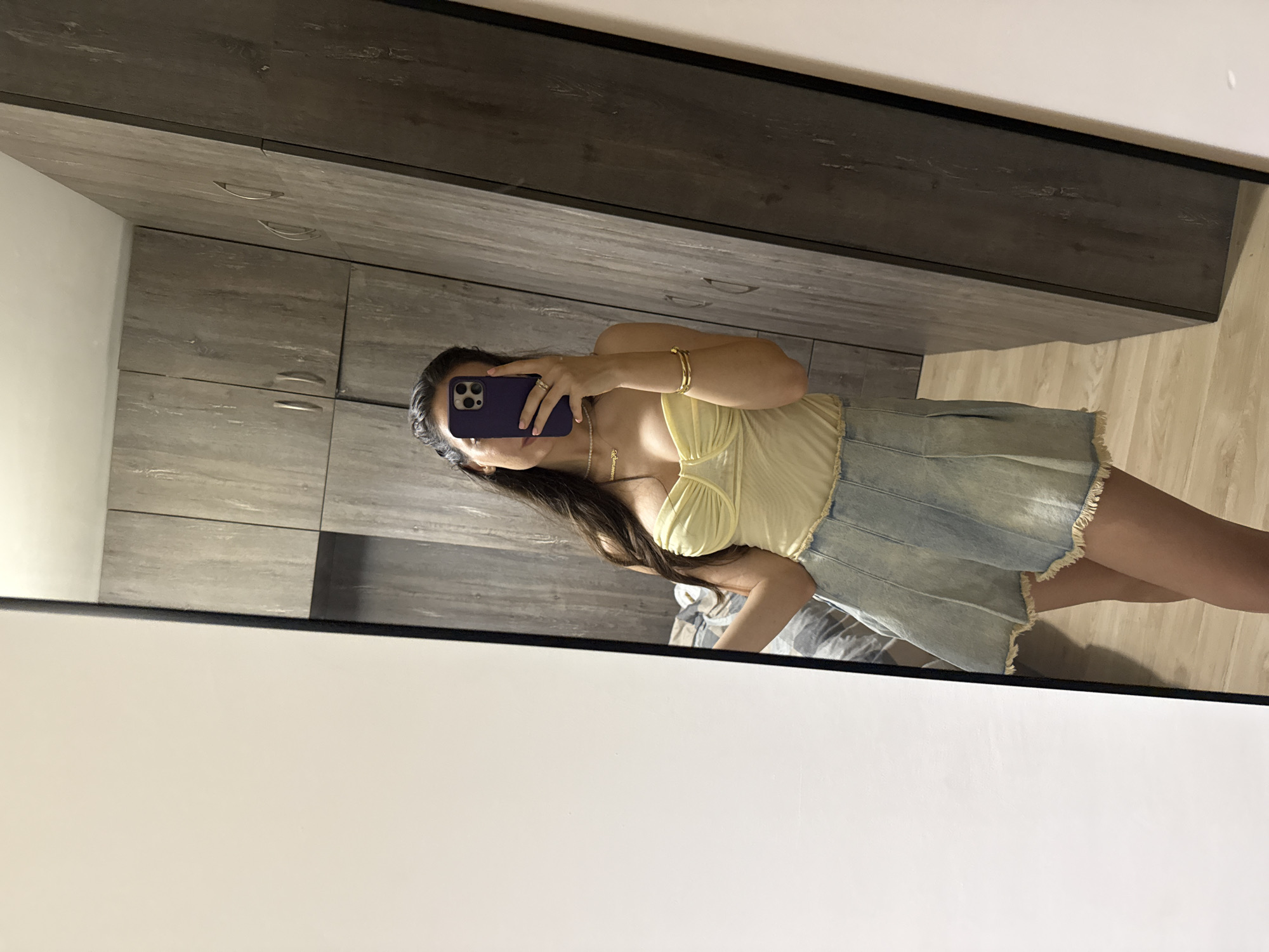 A woman takes a mirror selfie inside an apartment. The woman is wearing a yellow top and a light blue denim skirt. She is holding a dark-colored smartphone up to obscure her face. She is wearing jewelry including a bracelet and necklaces. The background shows a wardrobe, a bed, and a light-colored wooden floor. The location appears to be inside a modern apartment, but the details are insufficient to determine a specific location within Plovdiv, Bulgaria.