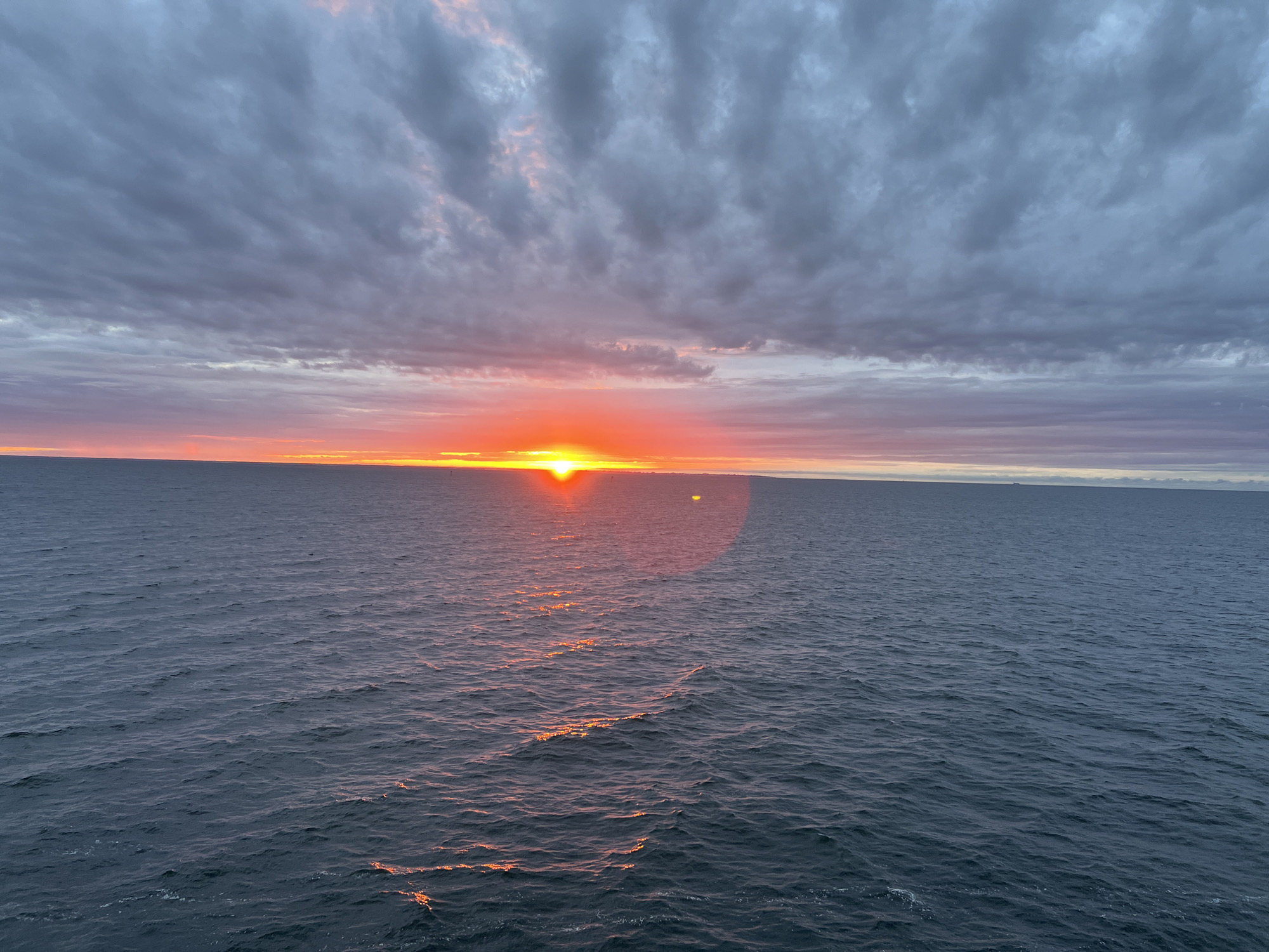 The image shows a sunset over the Baltic Sea, as viewed from Gdynia, Poland. The sky is filled with dramatic, grey clouds tinged with pink and orange from the setting sun. The sun itself is a bright, orange orb just above the horizon, casting a golden reflection on the dark, choppy water. The sea stretches out to the horizon, meeting a band of pale sky. There are no people, subjects, or objects clearly visible in the frame, creating a vast and tranquil seascape.