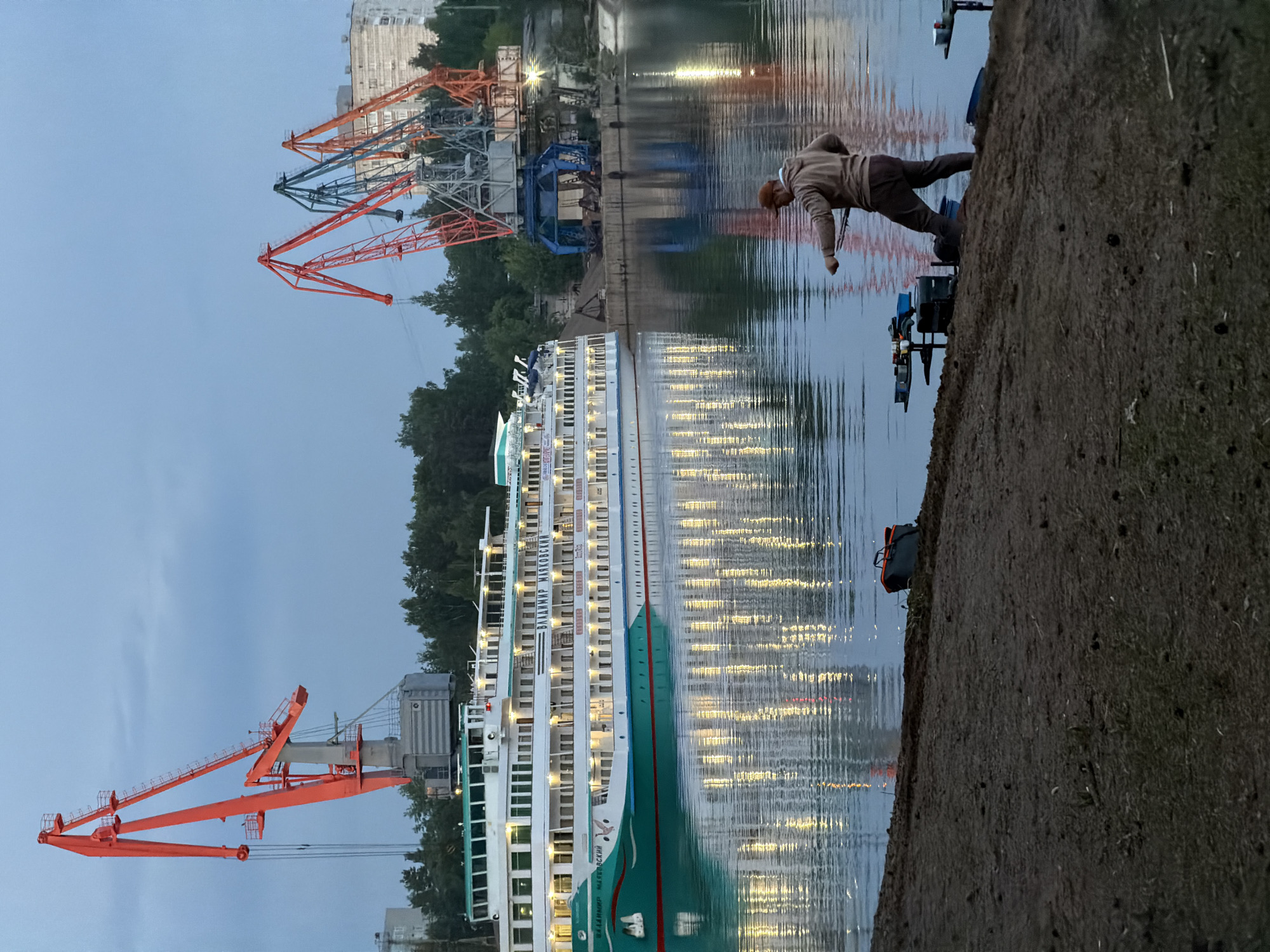 The image shows a man fishing from a concrete embankment in Chaikovsky, Russia. The city is recognizable by the cranes and cruise ship in the background. The man is leaning over the edge of the embankment with a fishing rod. Fishing equipment is visible on the embankment next to him. In the background, a large river cruise ship, the “Vladimir Mayakovsky,” is docked along the waterfront, and several port cranes are visible. The image appears to have been taken at dusk.