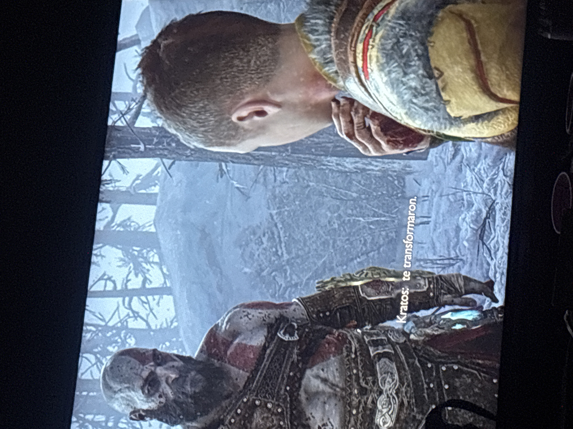 The image is a photograph of a screen displaying a scene from the video game "God of War." The primary subjects are the game's protagonists: Kratos and his son, Atreus. The scene is set in a desolate, snowy, and wintry forest, characterized by bare trees and snow-covered ground, indicative of a harsh northern environment.
In the lower half of the image, Kratos, the large, pale-skinned figure with prominent red markings and a stoic expression, is visible. He wears detailed battle armor, including a large gauntlet on his left arm. Kratos appears to be looking downwards, his head slightly tilted. In the upper half of the image, Atreus, a younger boy with short, dark hair, is featured. He is clad in fur-lined, warm attire suitable for the cold climate and is looking intently towards the left side of the frame, with his left hand slightly raised.
A dialogue box containing Spanish text is visible on the screen. The text reads: "Kratos: te transformaron." which translates to "Kratos: they transformed you." This indicates a moment of significant dialogue or a narrative event within the game.
The image is captured at an angle, showing some screen glare and pixelation, typical of a photograph taken of a digital display. The overall image is rotated 90 degrees clockwise, presenting the characters vertically instead of horizontally. The atmosphere is serious and contemplative, aligning with the dramatic tone of the "God of War" series. The photograph was taken in Buenos Aires, Argentina.