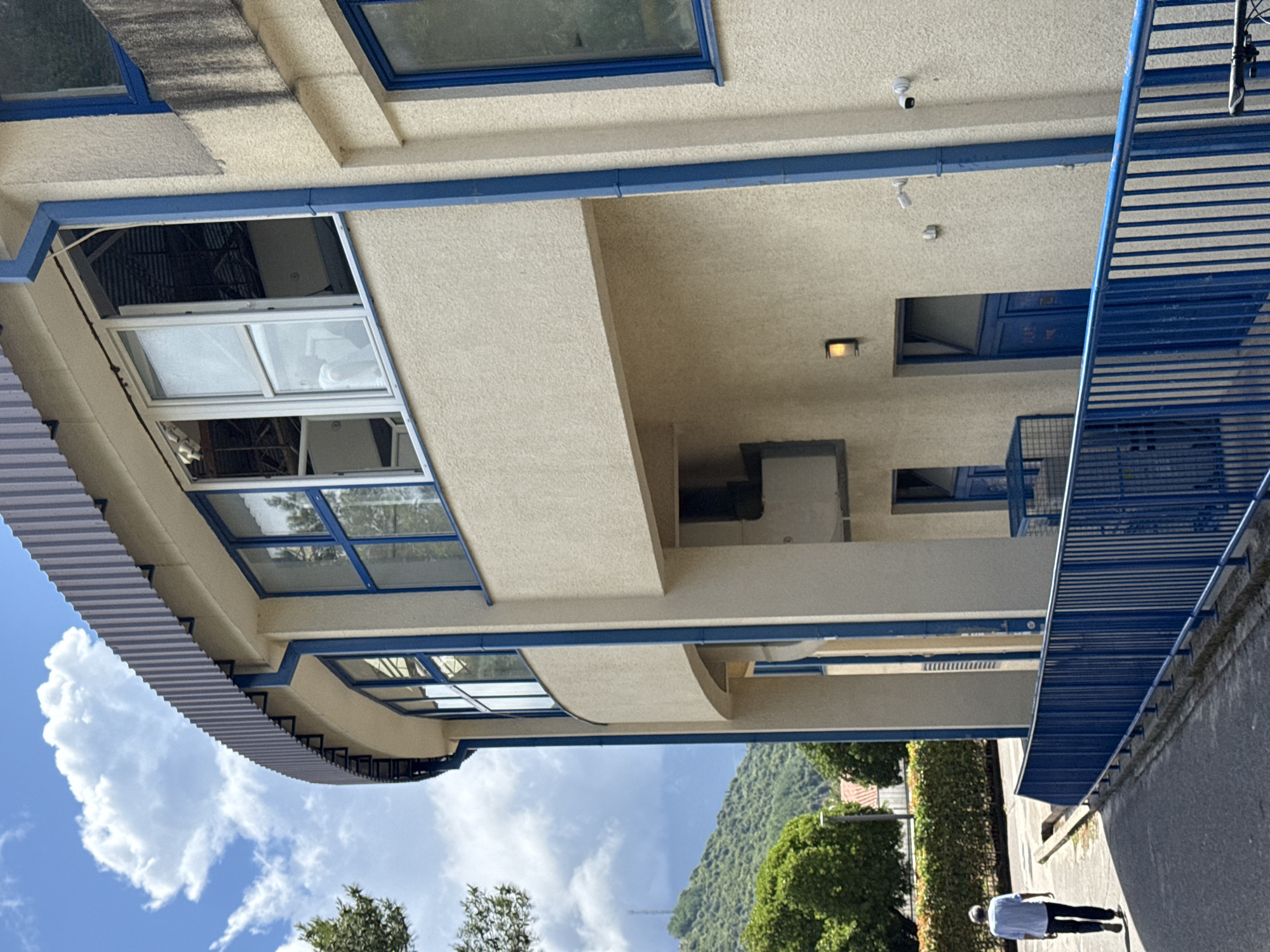 This image, rotated 90 degrees clockwise, depicts a modern, multi-story building under a bright blue sky with scattered white clouds. The building features a cream-colored, textured exterior and is characterized by prominent blue architectural elements, including window frames, trim lines, and a sturdy metal balustrade or railing on the right side.
A corrugated light blue awning extends from the building's left side. Several windows are visible, some with blue frames, while one large window section on an upper level is open, revealing a dark interior. Above this open window, a dark, streaky stain mars the building's facade. Curved architectural features are present beneath the awning.
On a paved area next to the building, a person wearing a light blue shirt and dark pants is walking away from the camera. In the background, a verdant, tree-covered mountain rises under the bright sky. Details on the building include security cameras and light fixtures mounted on the ceilings of sheltered areas. The scene takes place during the daytime, under sunny conditions. The location is Budva, Montenegro. No visible text is present in the image.
