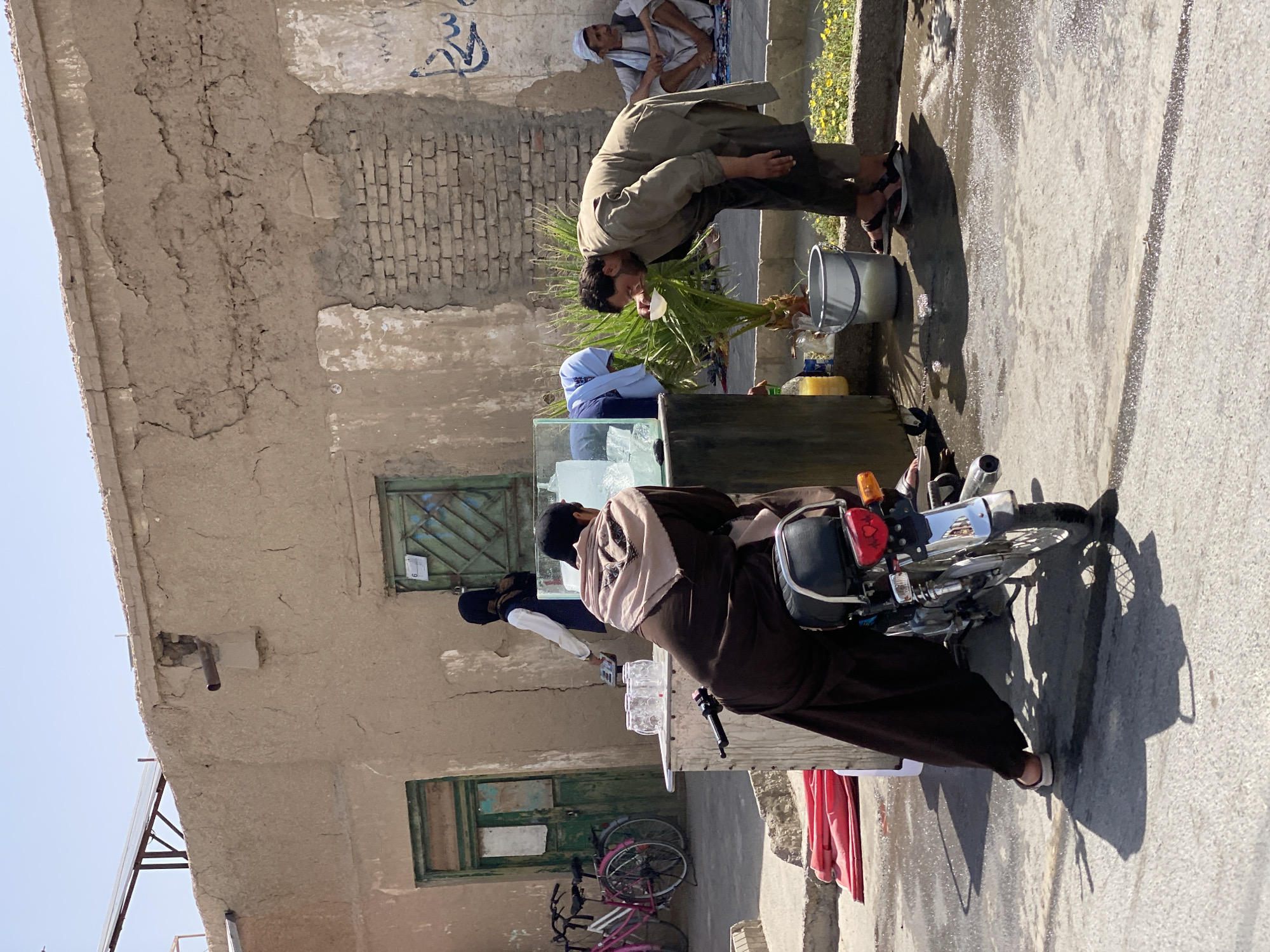 The image depicts an outdoor scene in a city, likely Kandahar, Afghanistan, characterized by a sun-drenched, dusty environment with aged, weathered buildings constructed from mud brick and stucco. In the foreground, a man dressed in a traditional brown shalwar kameez and a light brown shawl is seated on a motorcycle. To his right, another man in a muted green shalwar kameez is bending over a portable stand. This stand features a glass-fronted display case filled with ice and what appears to be drinks or condiments, and several empty glass cups are arranged on top. The man in green is drinking from a small white cup. Next to him, a palm plant is visible, and a silver bucket filled with liquid is on the ground. In the background, more buildings, some with brick details, are visible. A person in a dark hijab and white shirt is standing near a green door on the left, and two bicycles are parked nearby. Further to the right, two men are seated on the ground, one wearing a white turban. The ground is wet in places, suggesting recent activity. The overall impression is a snapshot of daily life in a warm, arid urban setting.