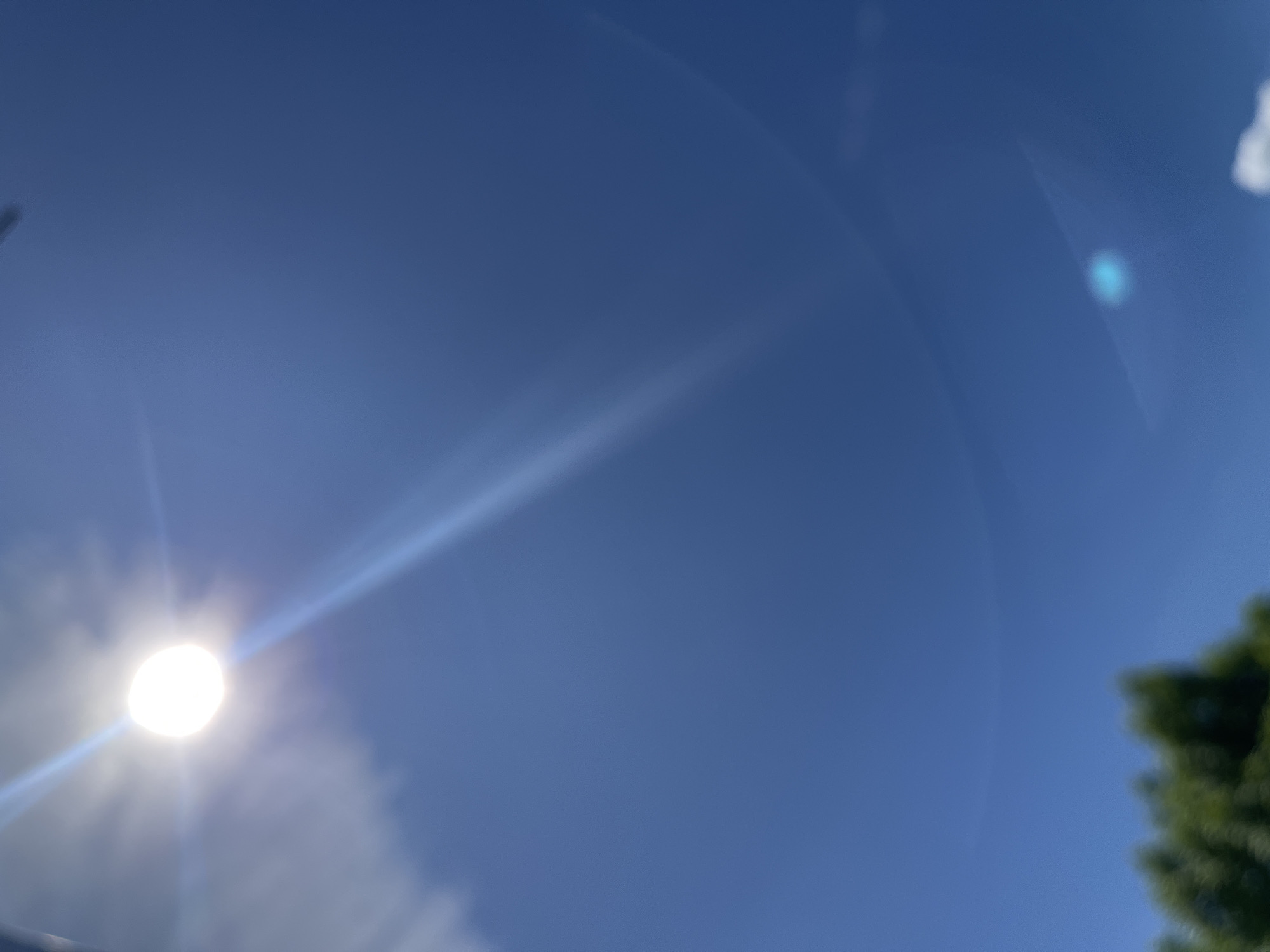 The image shows a view looking up at a bright sun in a blue sky. Lens flare and some clouds are visible around the sun. Some green foliage is present in the lower right corner of the frame.