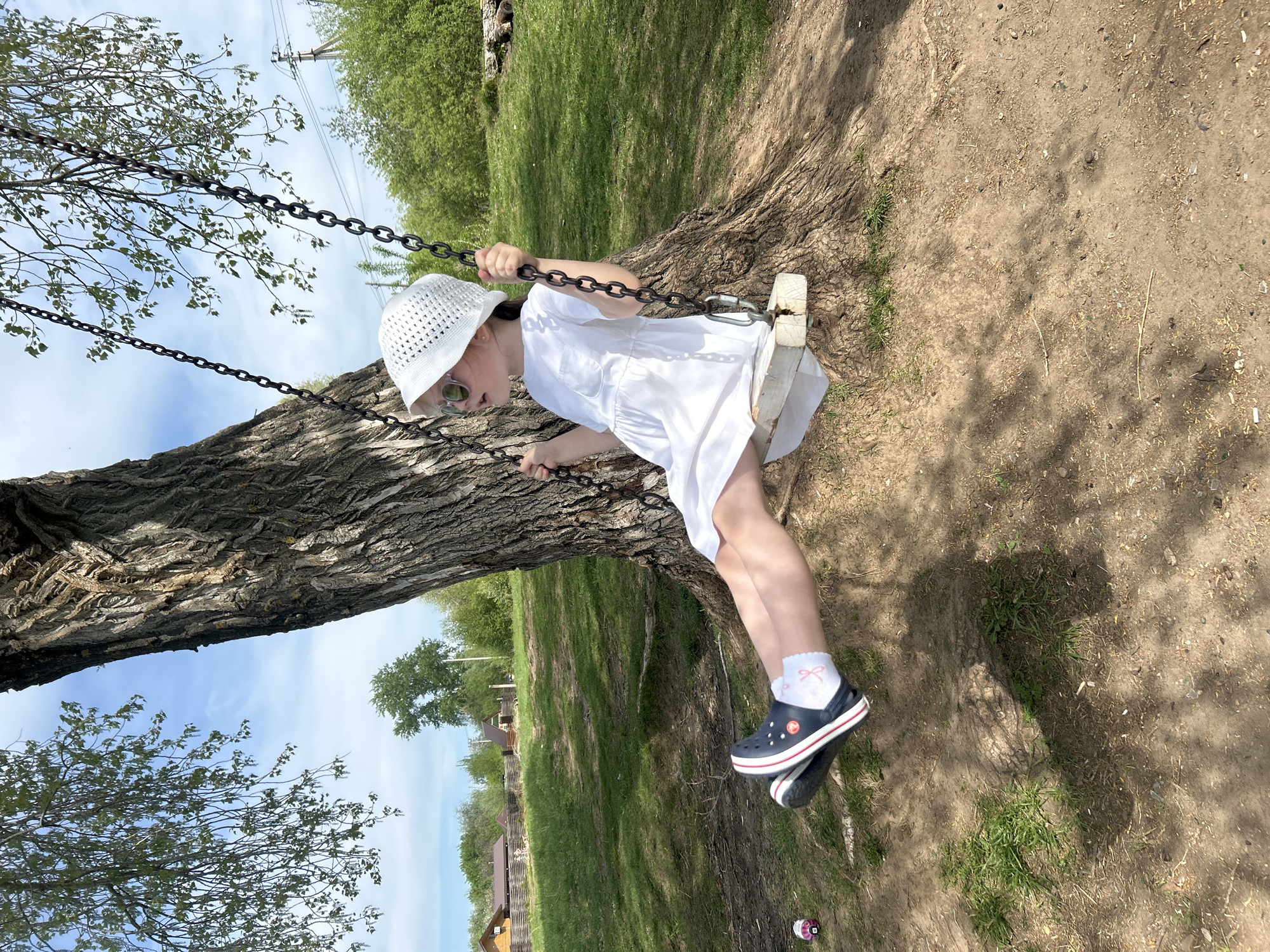 A young girl in a white dress and hat is on a swing suspended from a large tree. She is wearing sunglasses and navy blue Crocs with white socks. The ground is dirt and grass. In the background, there are green trees and houses. This photograph appears to be taken in a residential area, possibly in the city of Chaikovsky, Russia.