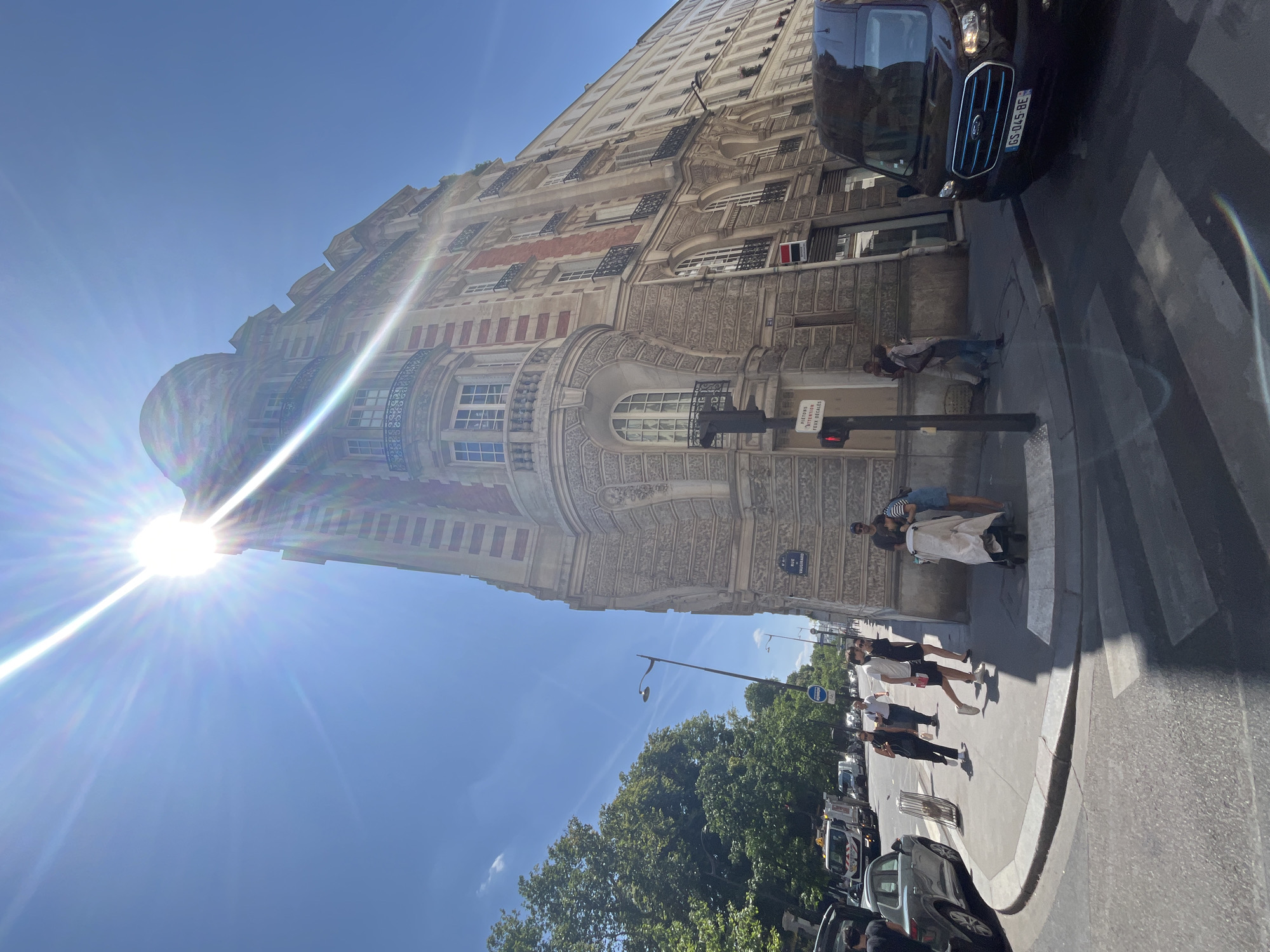 This is a street-level, wide-angle photograph taken on a sunny day in Paris, France, likely in the 8th arrondissement based on the architecture and street signs. The sun is intensely bright, creating a strong lens flare that dominates the upper left quadrant of the image.
The focal point of the image is a tall, ornate building with a rounded corner, constructed with red brick and stone detailing. The building features multiple stories with arched windows, balconies with wrought-iron railings, and decorative carvings. One sign on the building reads "SOC GENERALE". Another street sign indicates "RUE DE VAUGIRARD".
In the foreground, a black Ford van with a license plate starting with "GS 045 BE" is parked on the right side of the street, partially obscuring a crosswalk. Several pedestrians are visible on the sidewalk in front of the building. On the left side of the street, there are trees, a lamppost, a traffic light, and a street cleaning truck. People are walking and interacting on the sidewalk. One individual is pushing a stroller.
The sky is a clear, bright blue, and the overall scene suggests a bustling urban environment in Paris during daylight hours.