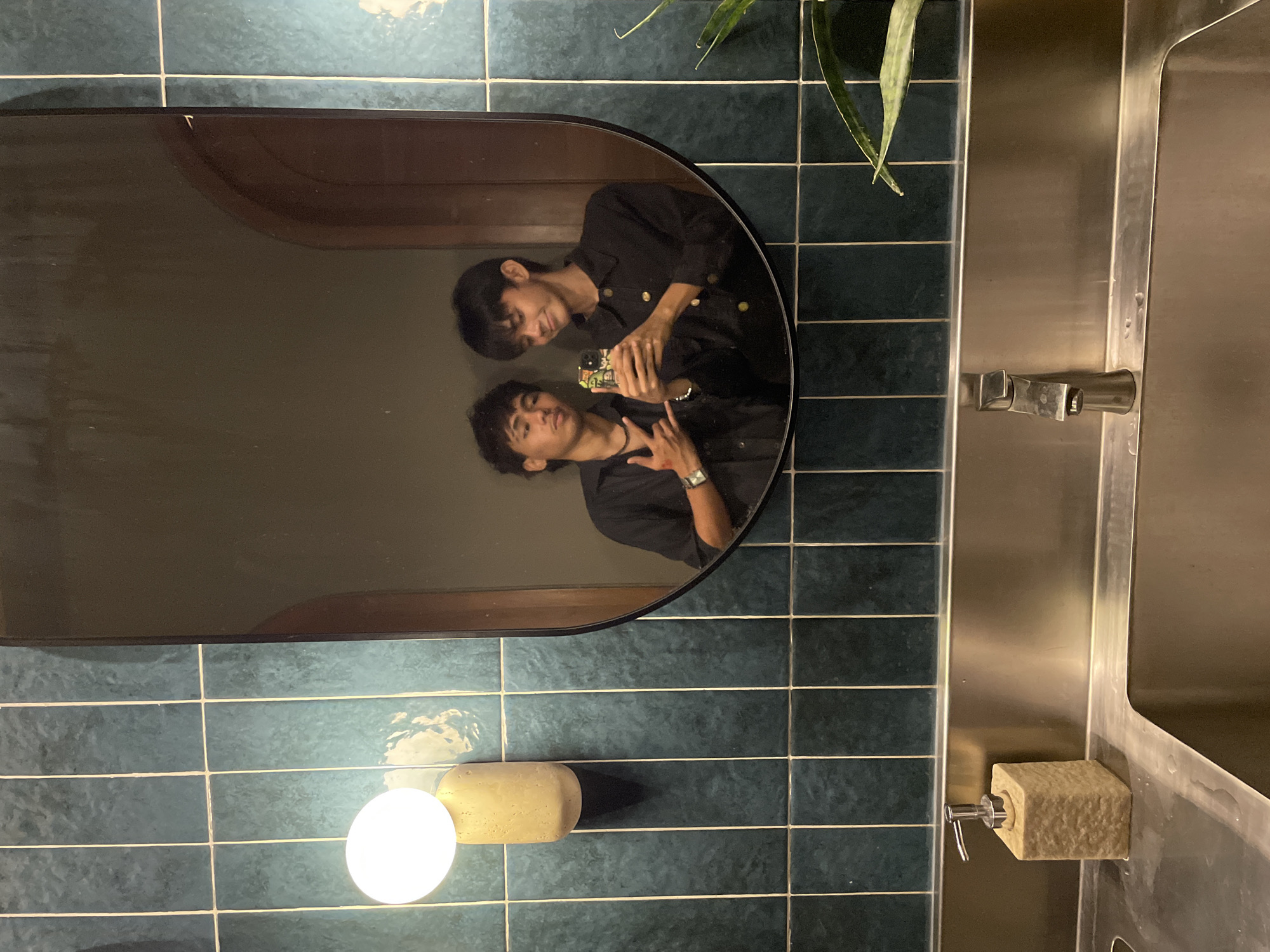 This is a mirror selfie taken in a restroom, likely at a restaurant or bar, in Davao City, Philippines. Two young males are pictured. The person on the left is wearing a black shirt and a necklace, with their right hand making a peace sign and their left hand outstretched. The person on the right is also wearing a black shirt and holding up a smartphone to take the picture.
The restroom features dark teal subway tiles on the wall, with a modern, oblong mirror hanging above a stainless steel sink. To the left of the mirror, a unique, cylindrical light fixture with a warm glow is mounted on the wall. A stone-textured soap dispenser sits on the left side of the sink, and a faucet is visible in the center. A Sansevieria plant, with its characteristic long, pointed leaves, is positioned to the right, partially obscuring the background. The overall ambiance suggests a contemporary and stylish establishment.