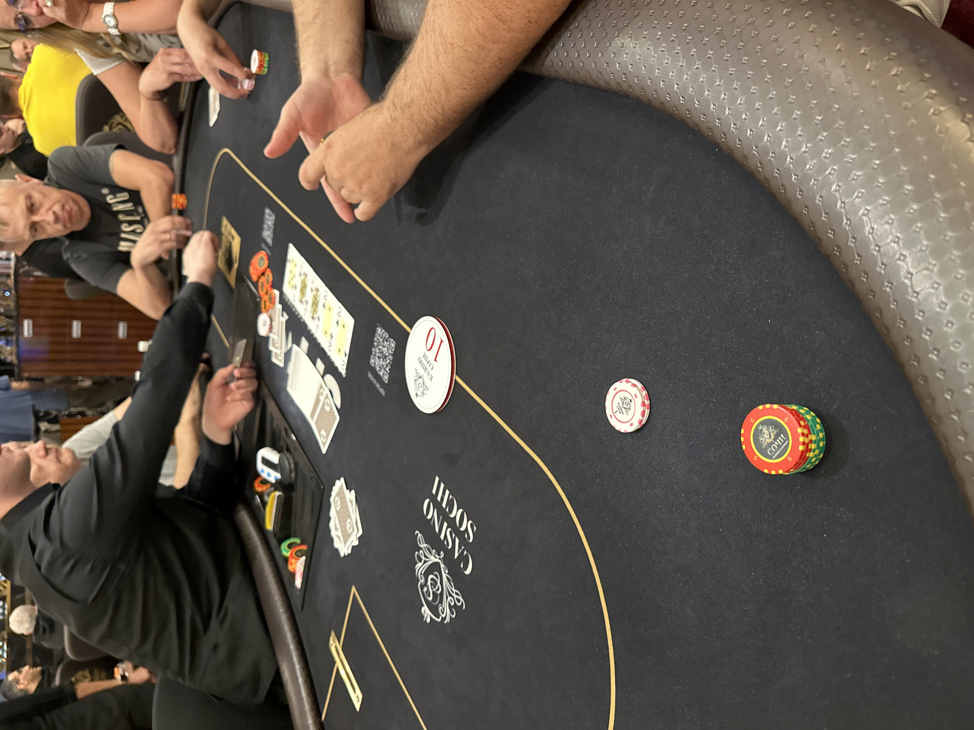 The image depicts a poker table at Casino Sochi in Estosadok, Russia. Several individuals are seated around the table, engaged in a poker game. The table is covered with a black felt cloth featuring the "CASINO SOCHI" logo. Various poker chips, cards, and other game-related paraphernalia are visible on the table. The lighting suggests an indoor setting, likely a casino floor. One person in the foreground, wearing a black shirt, is reaching towards the table. Another person, wearing a gray t-shirt with a graphic print, is looking down at the cards. A woman with glasses is also visible. The atmosphere appears focused and engaged in the game.