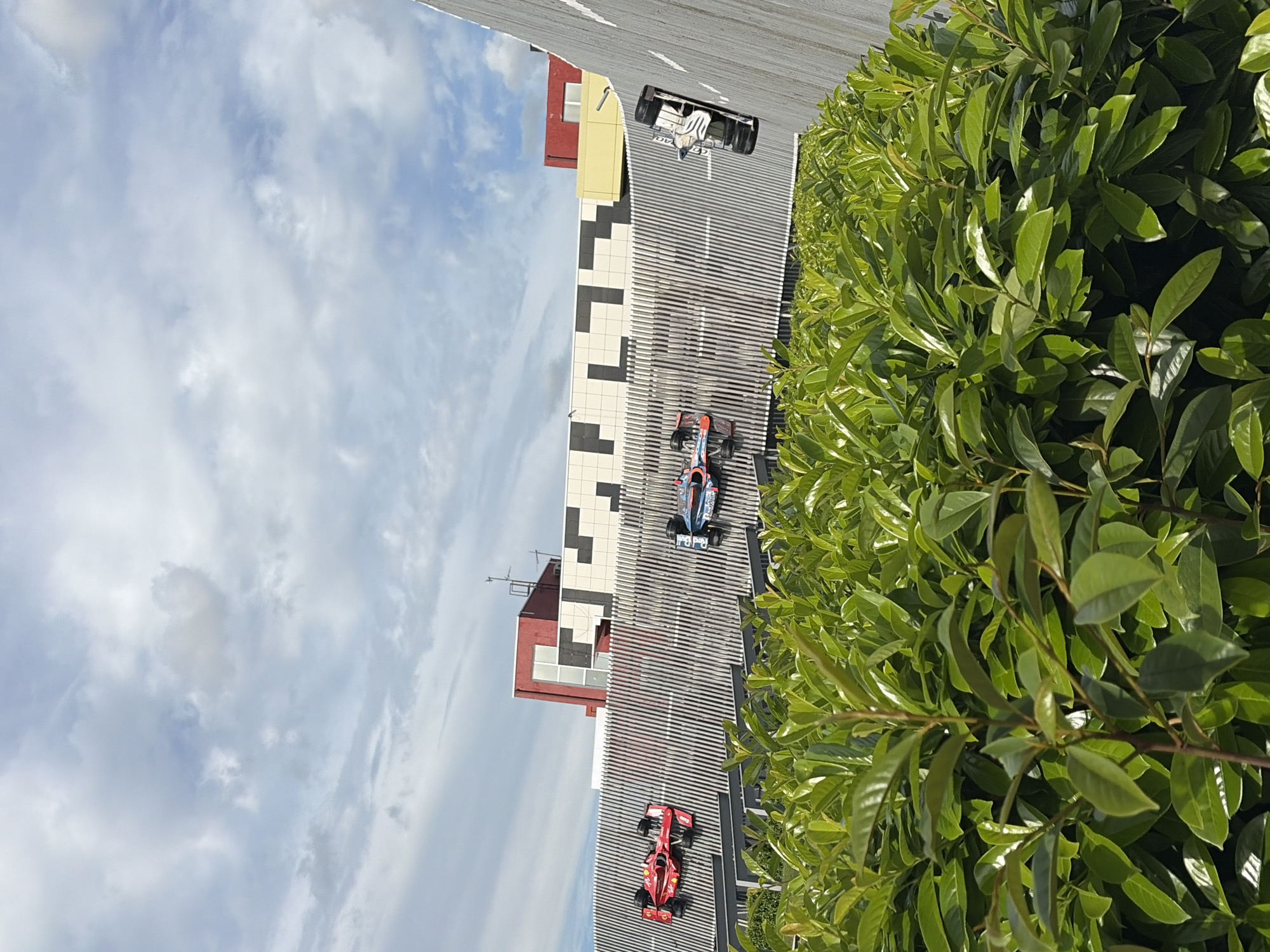 The image captures a section of a building facade in Anapa, Russia. The building features a modern design with alternating black and white square panels and red and yellow sections. Three toy Formula 1 racing cars are positioned on what appears to be a rooftop or balcony. The vehicles are red, blue, and white. Part of the frame is occupied by a bush with dense green foliage. The sky is visible in the background, with a mix of blue and white clouds.