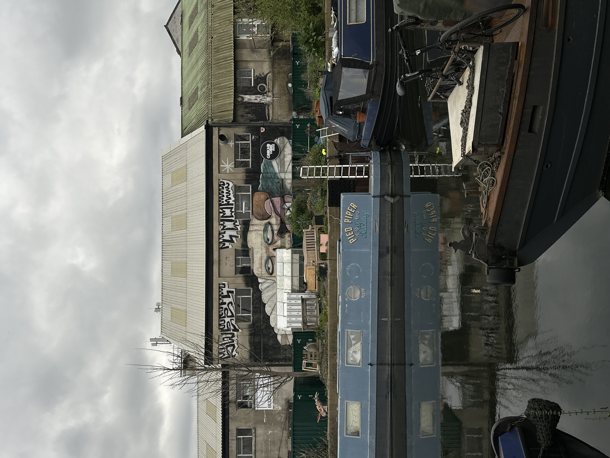 A riverside scene in London, United Kingdom, features a canal with reflections of buildings and boats. A multi-story industrial building is adorned with large graffiti artwork depicting a face. Adjacent to the building, a variety of boats are docked, including one prominently displaying the name "PIED PIPER". Various objects like ladders, bicycles, and personal belongings are visible on the boats and along the riverbank. The sky is overcast with heavy clouds. The overall environment suggests a blend of urban industrial elements and waterfront activity.