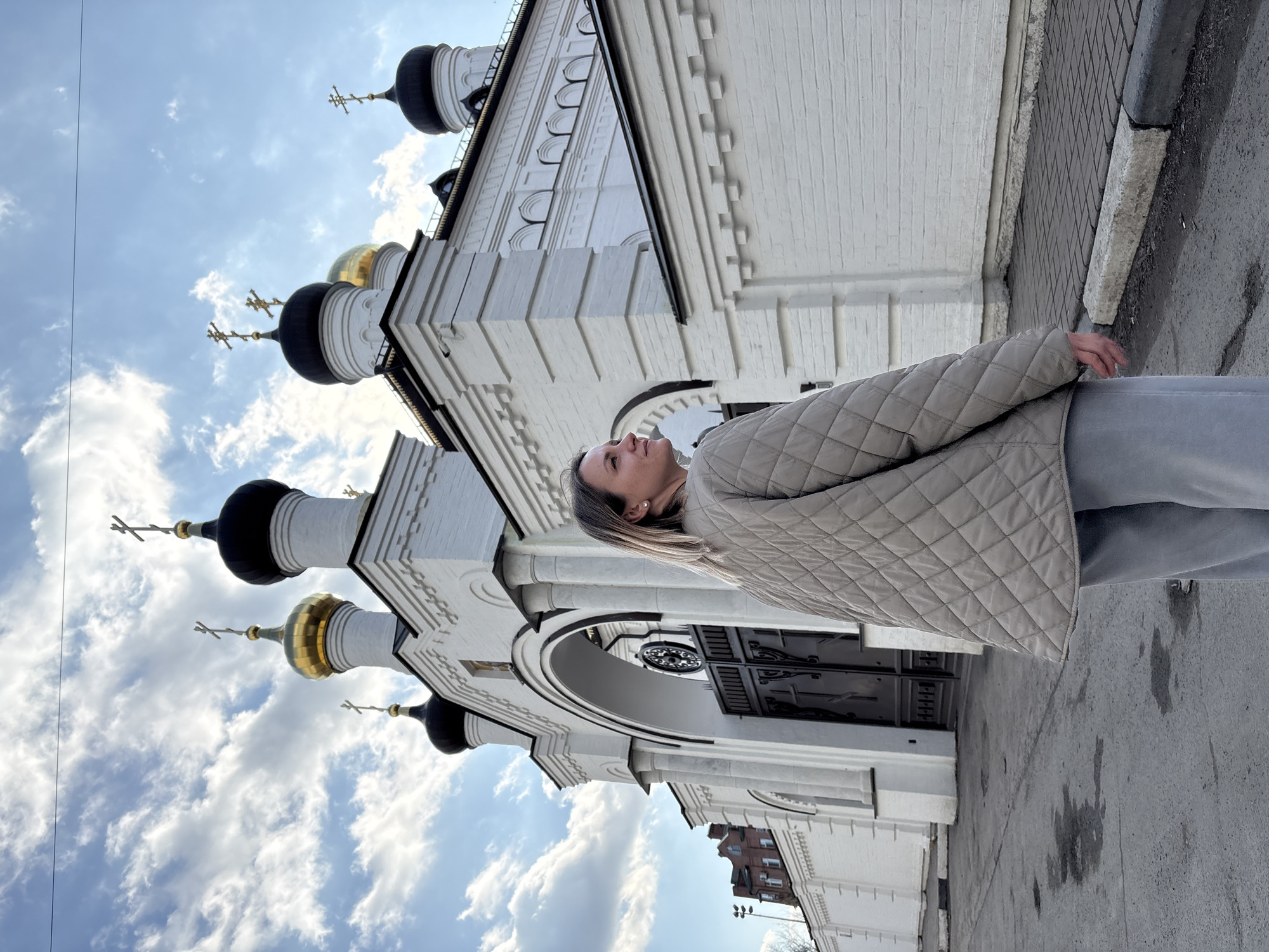 A young woman stands next to the Holy Resurrection Cathedral in Ulyanovsk, Russia. The church is white with several black and gold domes. The sky above is light blue with puffy white clouds. The woman is wearing a light beige jacket and gray pants. She is looking up at the church. The ground is gray and there is a sidewalk next to the building.