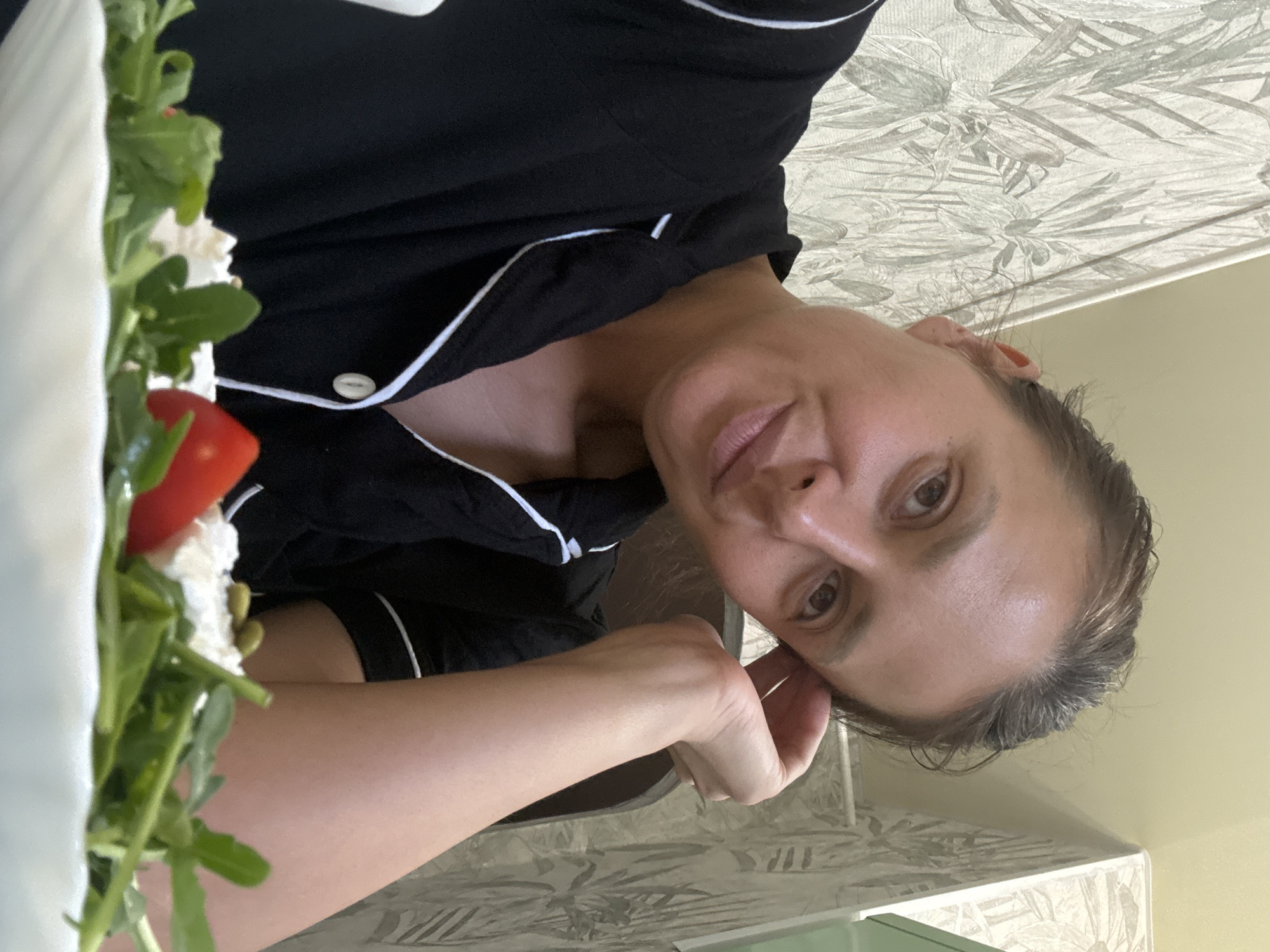 A woman with fair skin and brown eyes is seen at an extreme oblique angle, presumably indoors. She is wearing black pajamas with white piping. Her left hand is close to her face. To the left of the frame, a plate is partially visible with salad, including tomatoes, white cheese, and greens. The background includes floral wallpaper and a light yellow wall. The location is Chaikovsky, Russia.