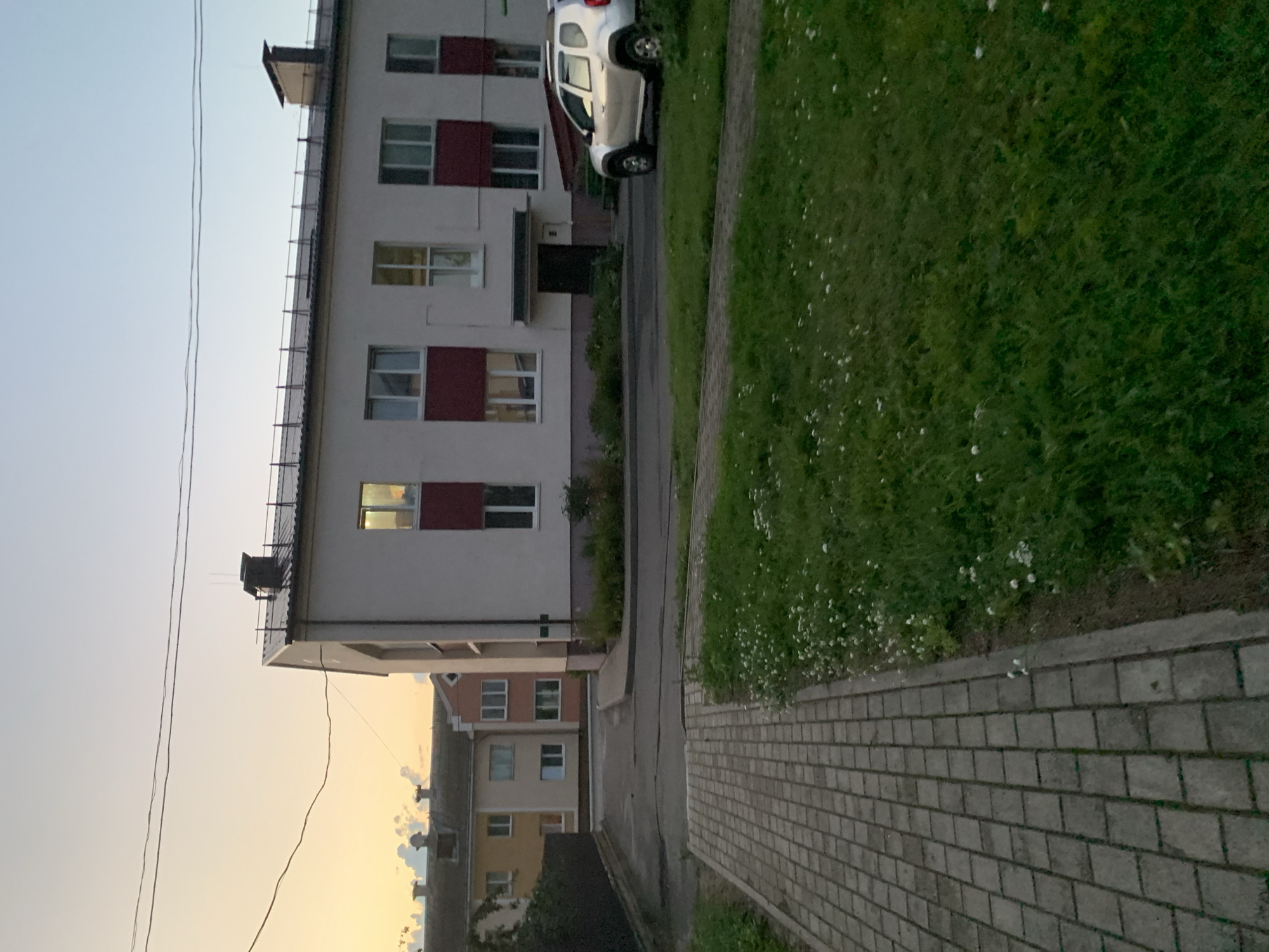 An outdoor, eye-level shot captures a residential street in Kapyĺ, Belarus, during dusk. The sky is painted with soft hues of orange and yellow, transitioning into a pale blue overhead. Power lines stretch horizontally across the sky.
Dominating the right side of the frame is a modern, two-story building with a white facade and dark red accents around its numerous windows. The building features a dark, flat roof with visible railings and a chimney. A silver SUV is parked on the asphalt road in front of this building.
To the left of the main building, further down the street, are two other residential buildings. The closer one is a two-story structure with a yellowish-orange facade, and the one behind it has a lighter, peach-colored exterior. Both exhibit multiple windows. A dark fence lines the left edge of the image, partially obscuring the view of these buildings.
A paved walkway made of grey interlocking bricks leads from the bottom left corner of the image into the scene, following the curve of the road. Adjacent to the walkway and extending across the foreground and mid-ground is a patch of green grass dotted with small white wildflowers. The road itself is asphalt, showing signs of wear and cracks. The overall impression is of a quiet, suburban neighborhood at the end of the day.