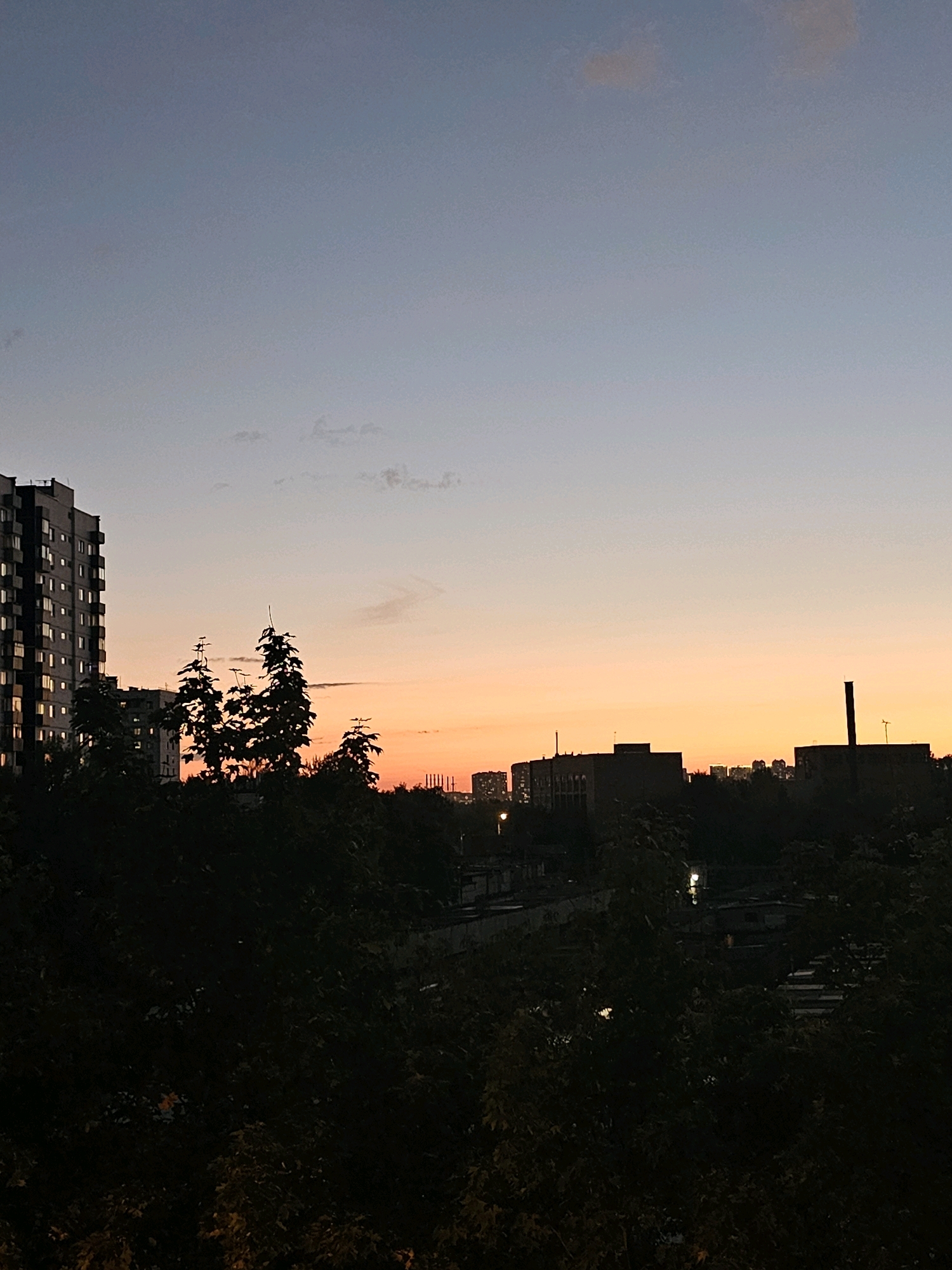 This image captures a twilight scene in Khimki, Russia, featuring a silhouette of the city skyline against a gradient sky. In the foreground, dark masses of trees obscure the lower half of the frame. To the left, a tall, multi-story building with numerous illuminated windows stands out against the dusky sky. Behind it, more apartment buildings are visible, also silhouetted. In the middle ground and to the right, the skyline is punctuated by industrial-looking structures, including what appears to be a factory or processing plant with smokestacks. Faint lights from distant buildings are scattered across the horizon. The sky transitions from a pale blue at the top to soft hues of orange and pink near the horizon, indicating the end of daylight. No people or discernible actions are present in the image.