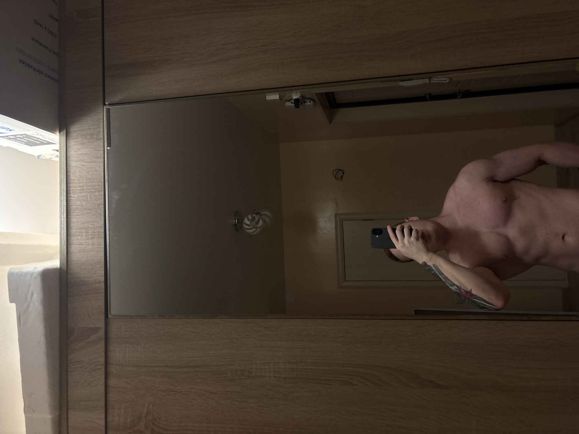A shirtless man is reflected in a mirror, holding a phone in his left hand and appearing to take a picture of himself. The man has a tattoo on his left arm. The mirror is mounted on a wooden surface, possibly a cabinet or wall. In the background reflected in the mirror is a bathroom with a light fixture, light-colored walls, and a door frame. The location, Chaikovsky, Russia, cannot be verified from the image.