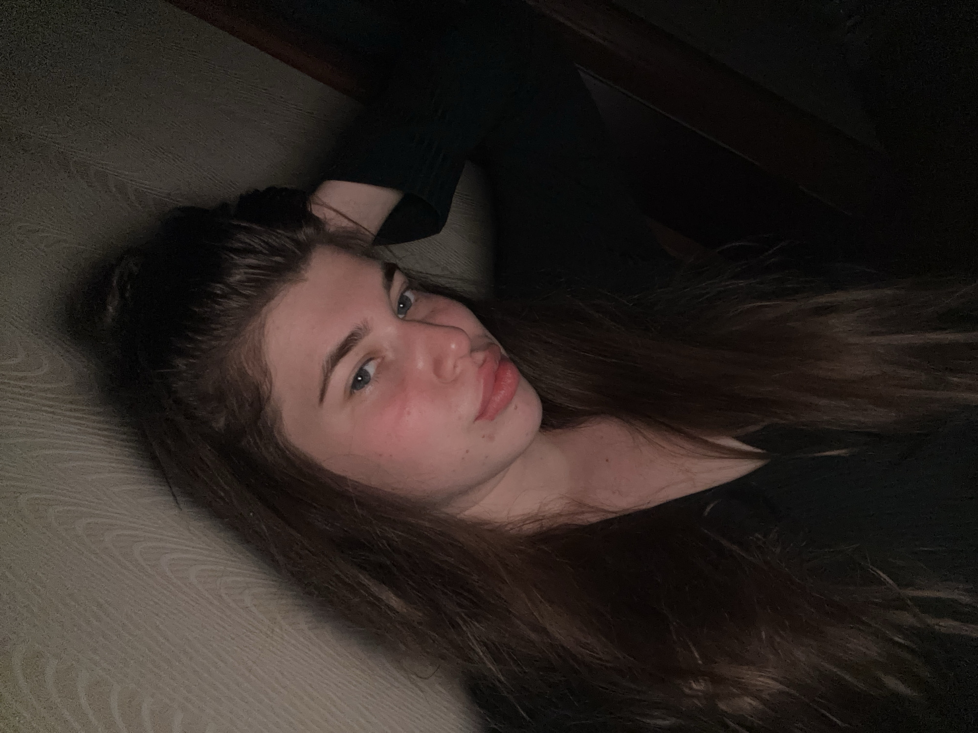 A young woman lies on a patterned cushion or upholstered surface. She has long brown hair, light eyes, and rosy cheeks. She is wearing a dark-colored shirt. The setting appears to be indoors, dimly lit. There is no indication of the city of Мещёры, Russia.